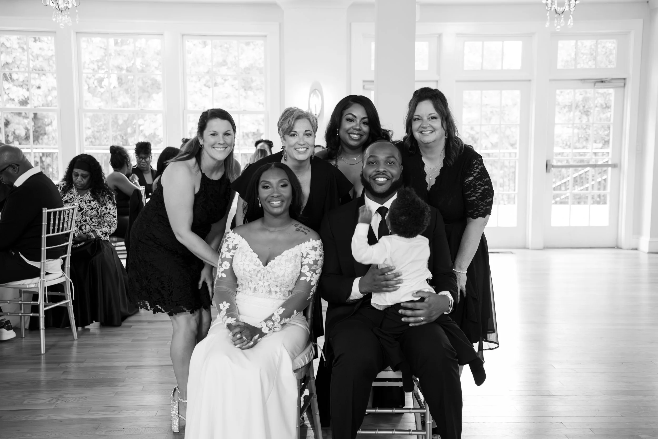 A group of seven people, including a bride and groom, smiling and posing for a photo at a celebration in a bright room with large windows. Photo & Video by Will Locke Wedding Photography at the Woman's Club of Portsmouth in Virginia. 