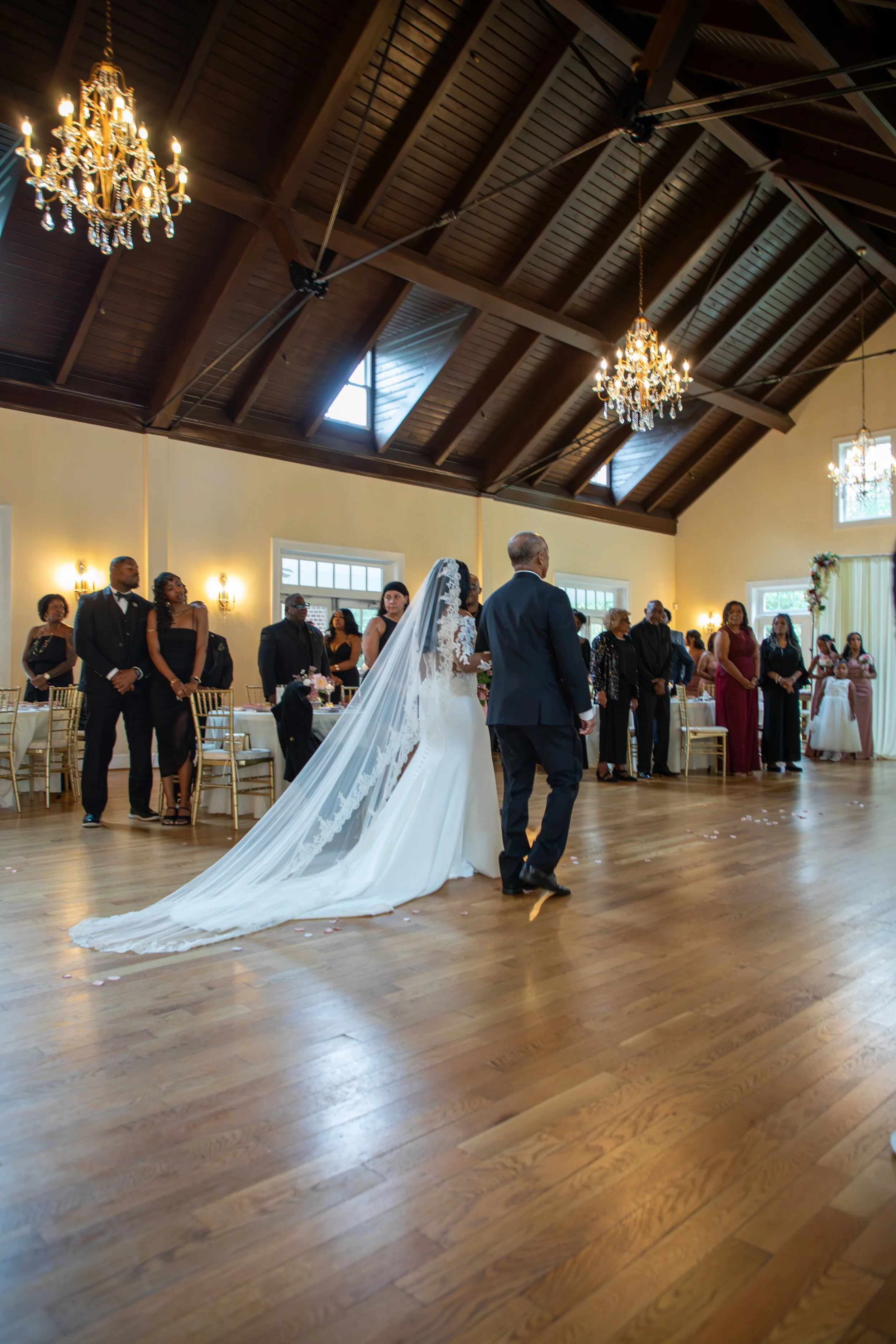 Wedding Photography by Will Locke. Bride walking down the aisle with a man, likely her father, at a wedding reception in a decorated hall with chandeliers and guests watching.