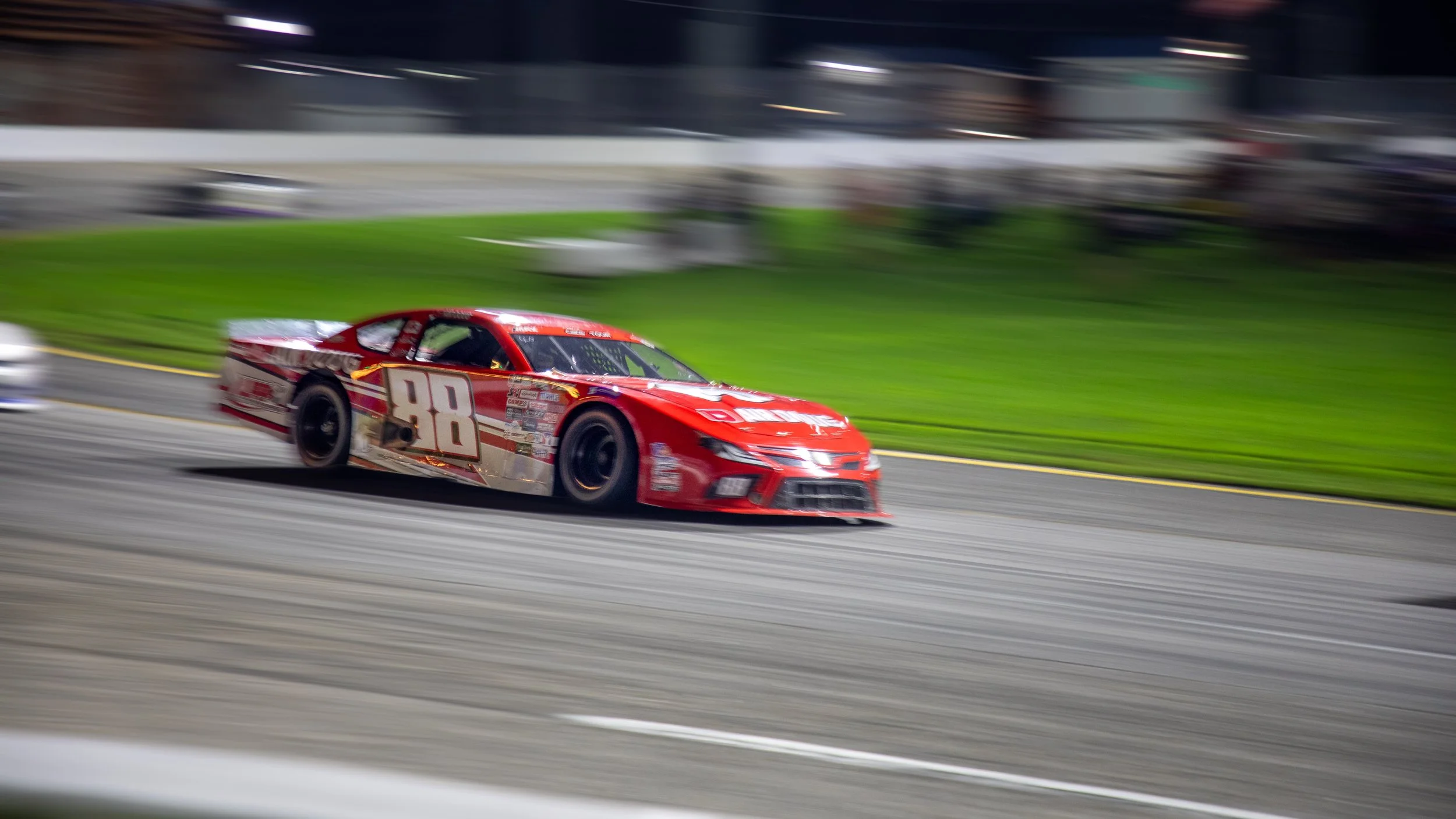 A red race car numbered 98 speeding on a racetrack at night, with a blurred background and track.