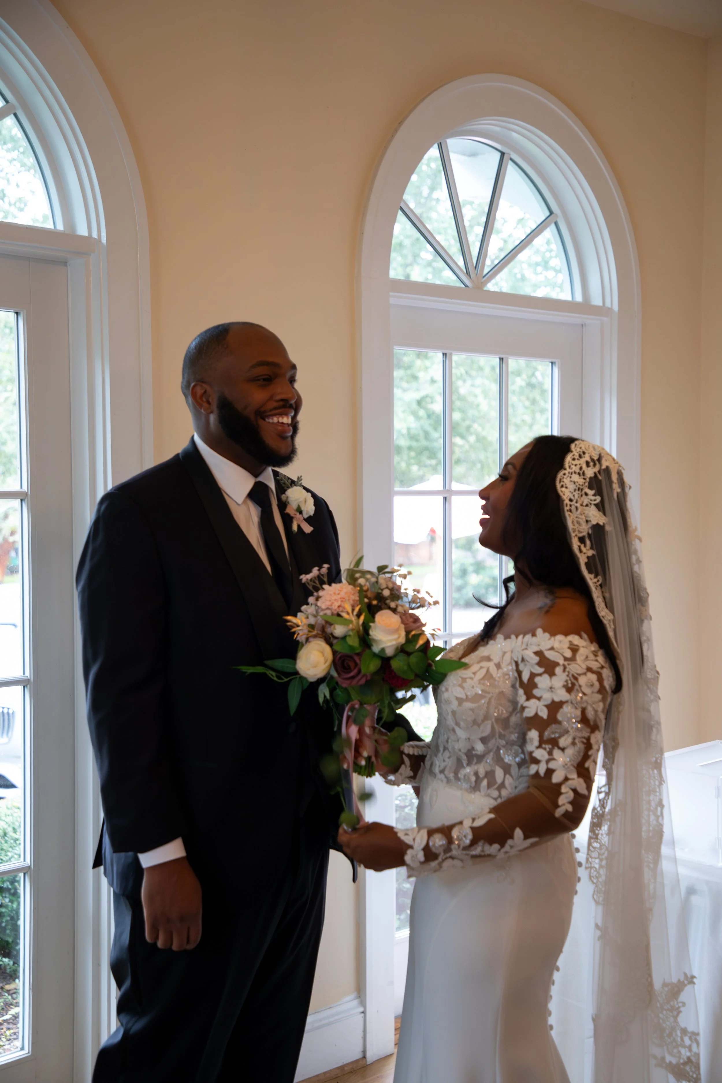 A bride and groom standing indoors near large windows, smiling at each other during their wedding ceremony. The bride holds a bouquet of flowers, and she is wearing a lace wedding dress with a veil. Wedding Photography by Will Locke. 