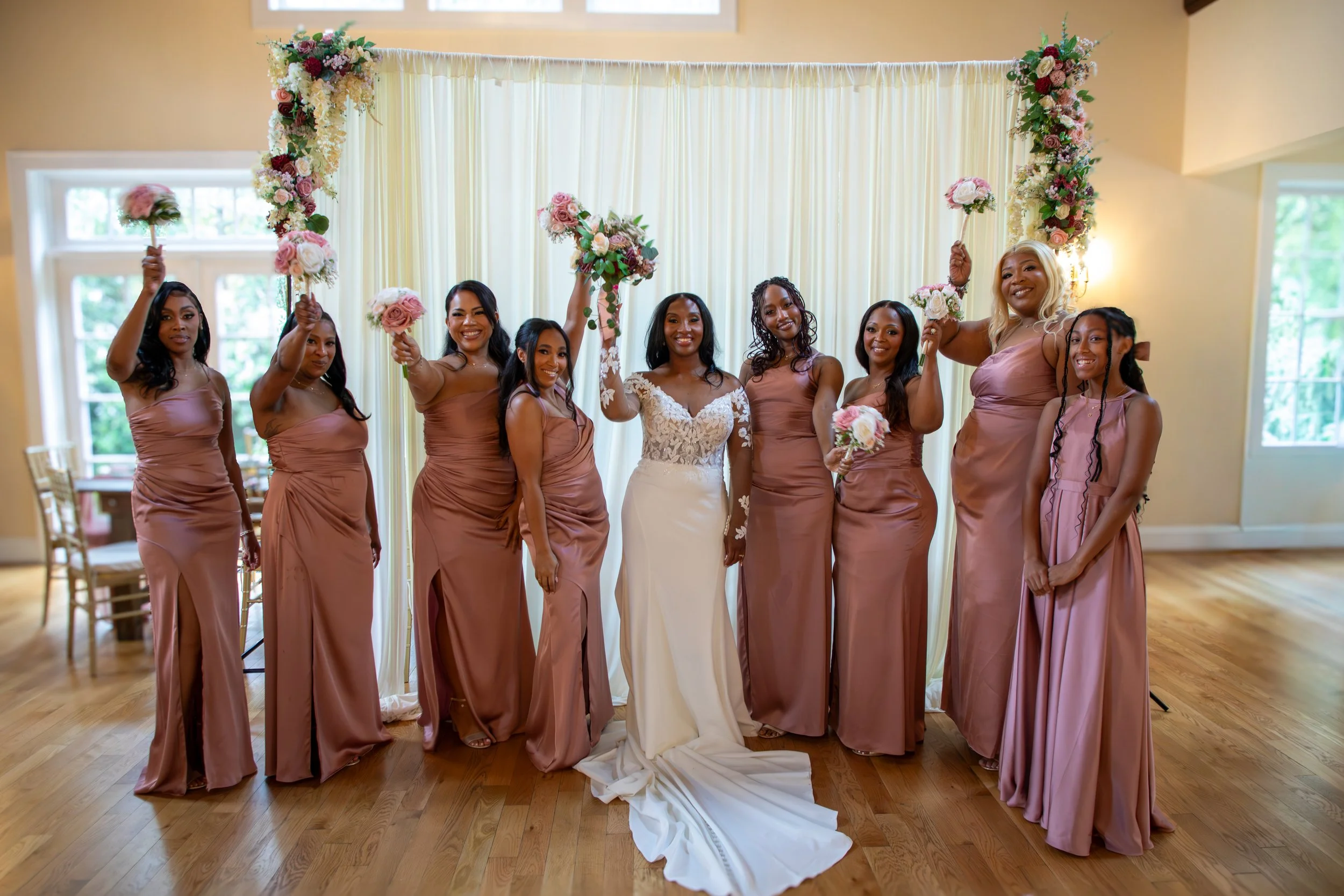 Wedding Photography by Will Locke. A bride in a white wedding dress stands in the center with seven bridesmaids in pink dresses, holding pink and white bouquets, smiling at a wedding reception.