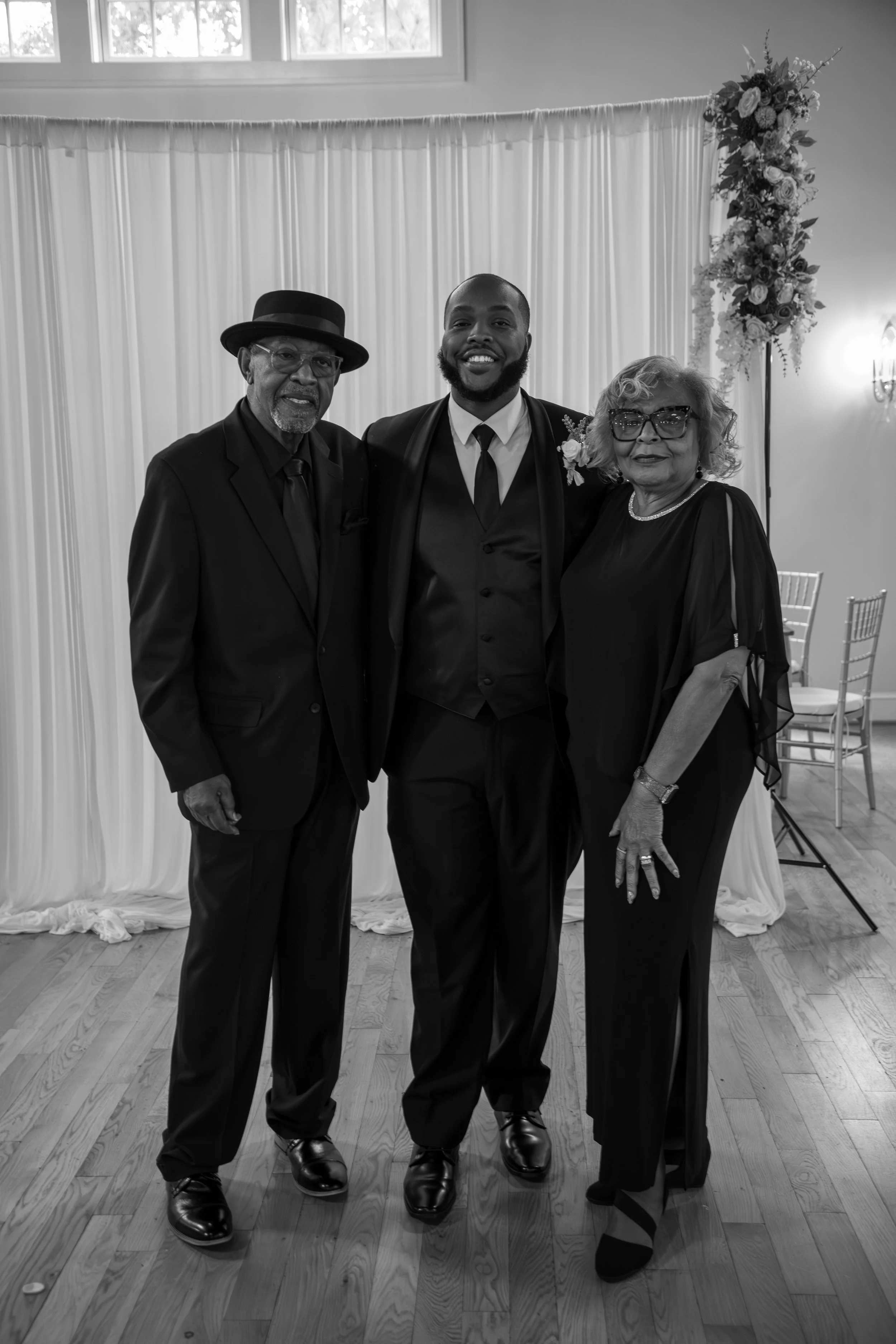 Photo & Video by Will Locke Wedding Photography at the Woman's Club of Portsmouth. A smiling young man in a tuxedo standing between an older man and woman, all at a formal event with a decorated backdrop, wooden floor, and chair in the background.