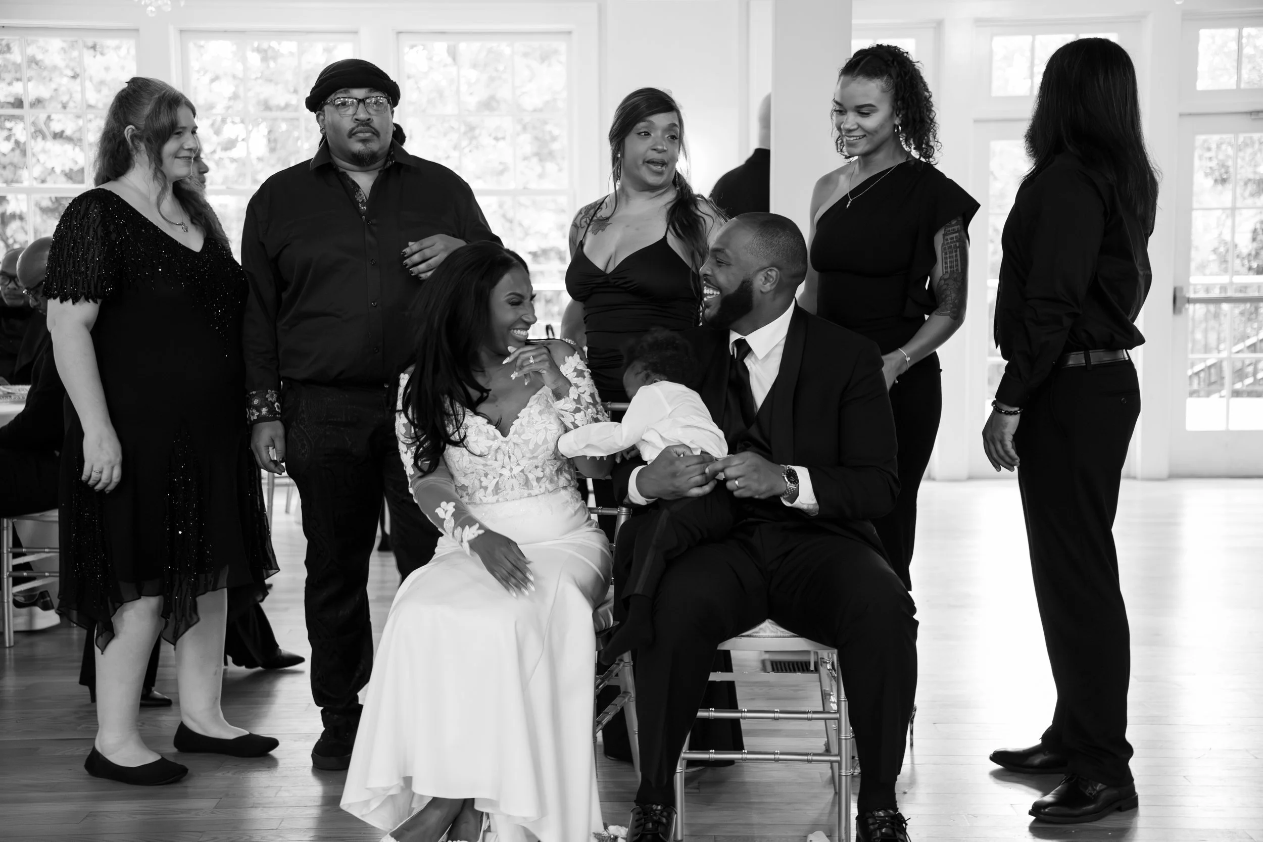 Photo & Video by Will Locke Wedding Photography at the Woman's Club of Portsmouth in Virginia. A group of seven people celebrating a joyful moment at a wedding, with a woman in a wedding dress and a man in formal attire holding a child.