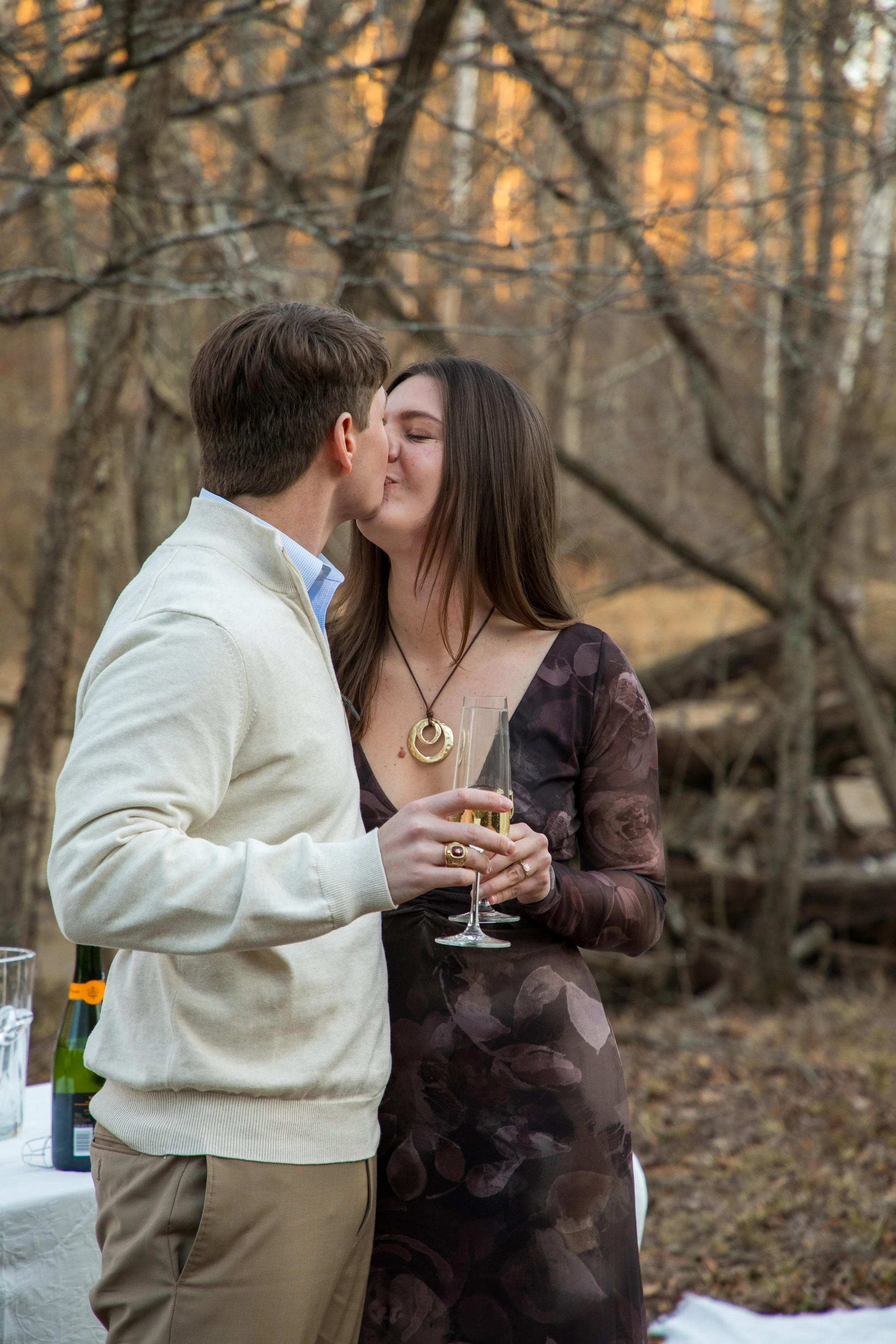 Engagement Photography by Will Locke near Richmond, VA in Montpelier. A man and woman kissing while holding champagne glasses outdoors during sunset, with a table of flowers nearby.