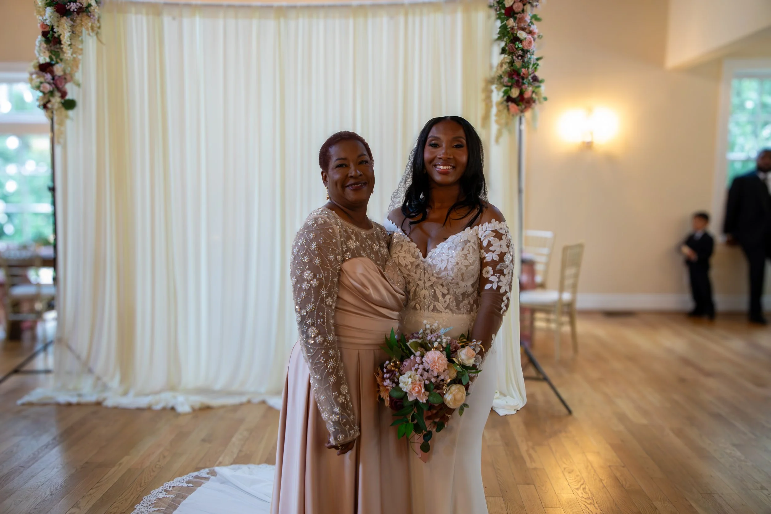 Two women smiling at a wedding, one in a lavender gown with long sleeves and the other in a white lace wedding dress. Photo & Video by Will Locke Wedding Photography at the Woman's Club of Portsmouth in Virginia. 