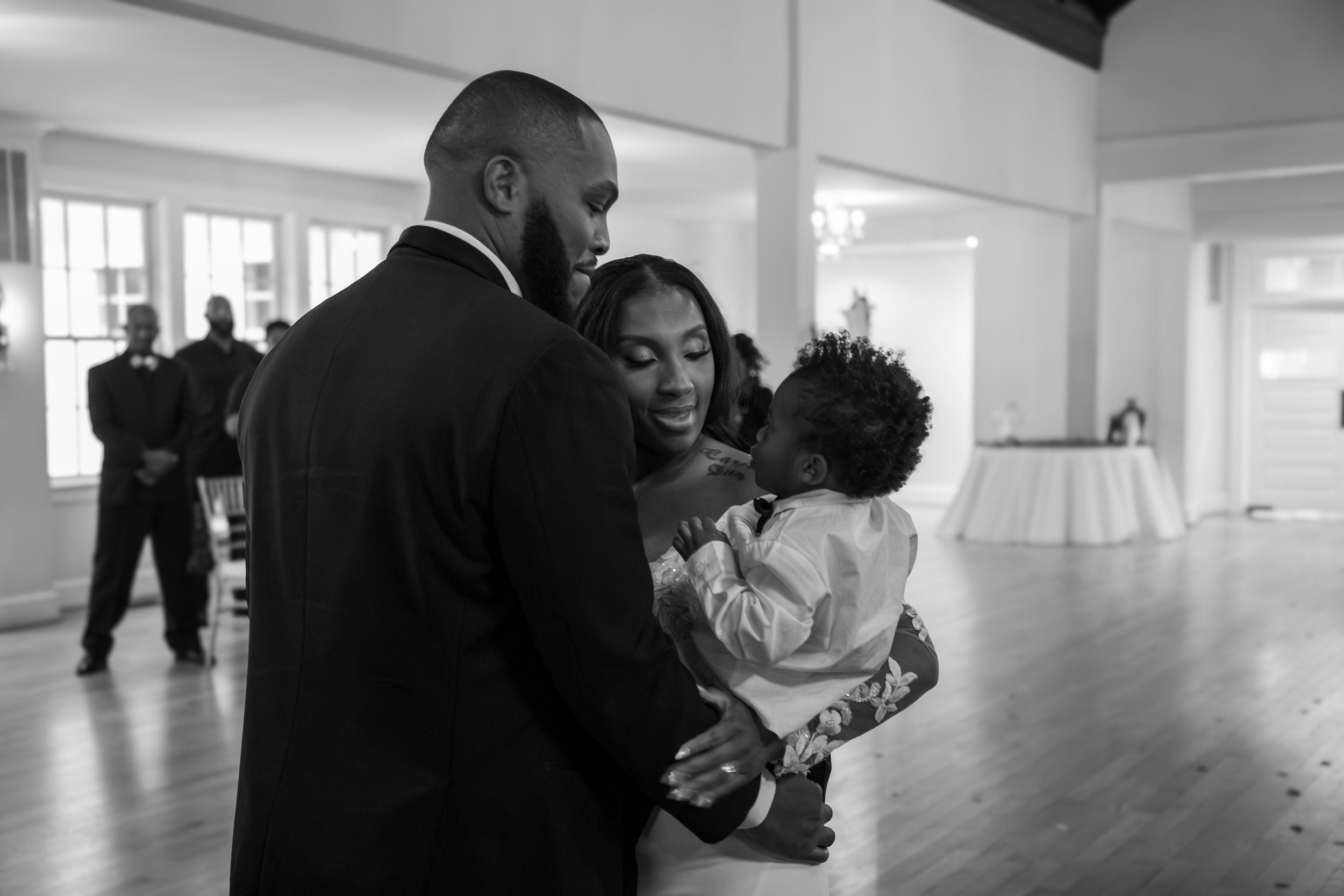 A black-and-white photo of a family at a wedding, with a man in a suit holding a young child and a woman in a dress. Photo & Video by Will Locke Wedding Photography at the Woman's Club of Portsmouth in Virginia. 