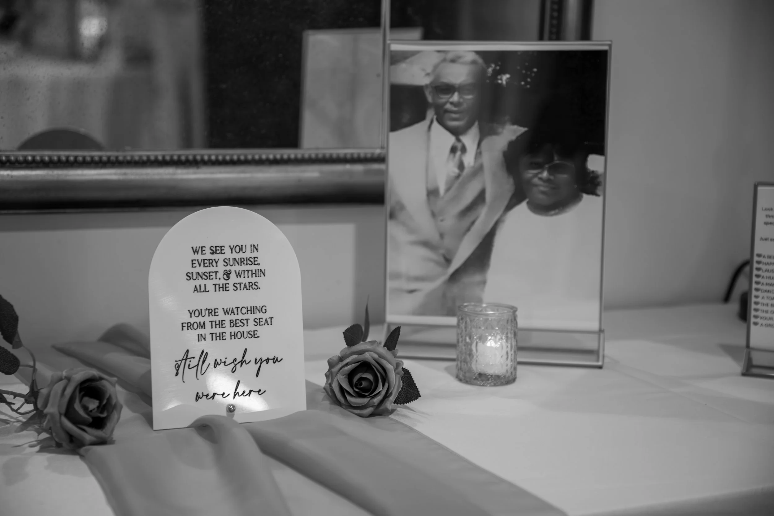 Wedding Photography by Will Locke. A memorial display with a framed photograph of two people, a candle, and artificial roses on a table. A card with a heartfelt message is also visible.