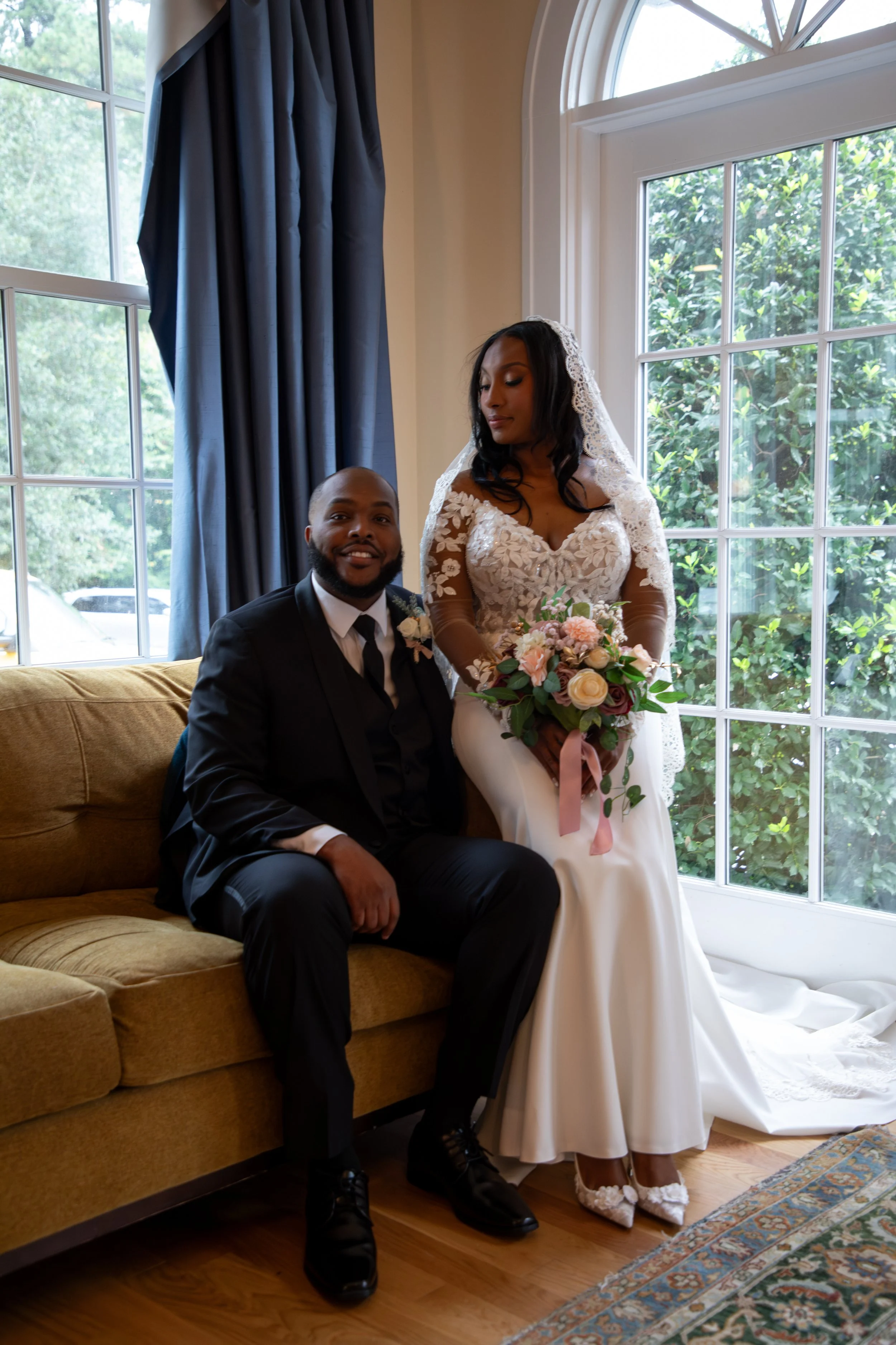 Photo & Video by Will Locke Wedding Photography at the Woman's Club of Portsmouth in Virginia. A bride holding a bouquet of flowers stands beside her seated groom in a room with large windows and blue curtains.