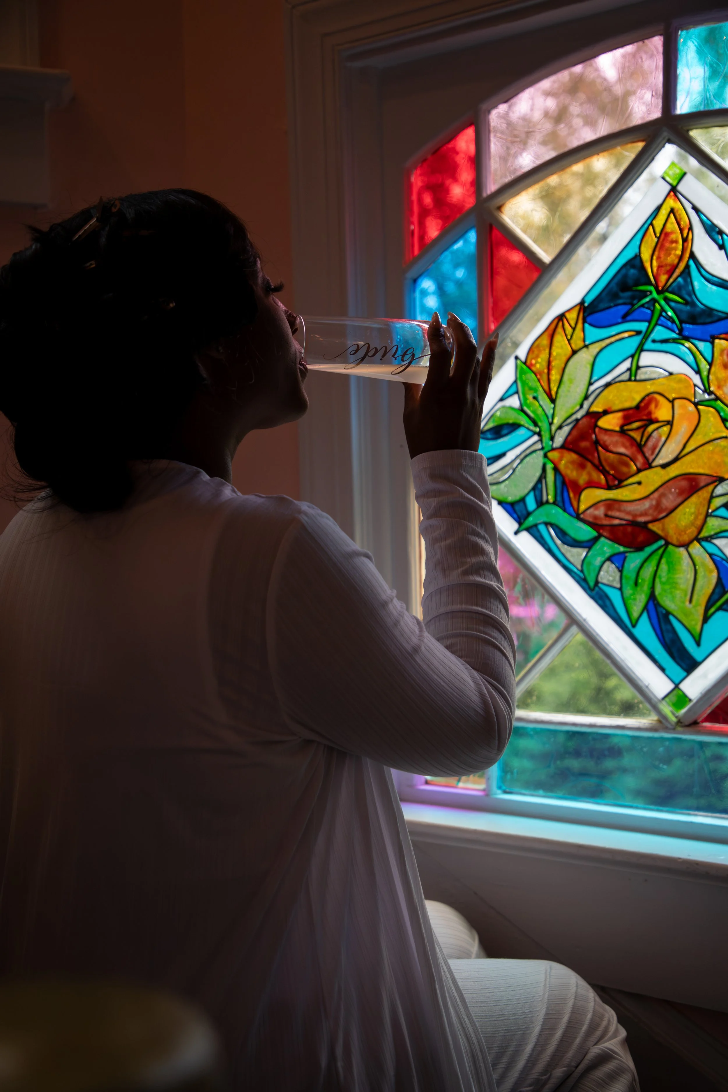 Photo & Video by Will Locke Wedding Photography at the Bridgerton House in Portsmouth, VA. A woman with dark hair in a bun, wearing a white long-sleeved shirt, drinks from a glass of champagne before her big day.