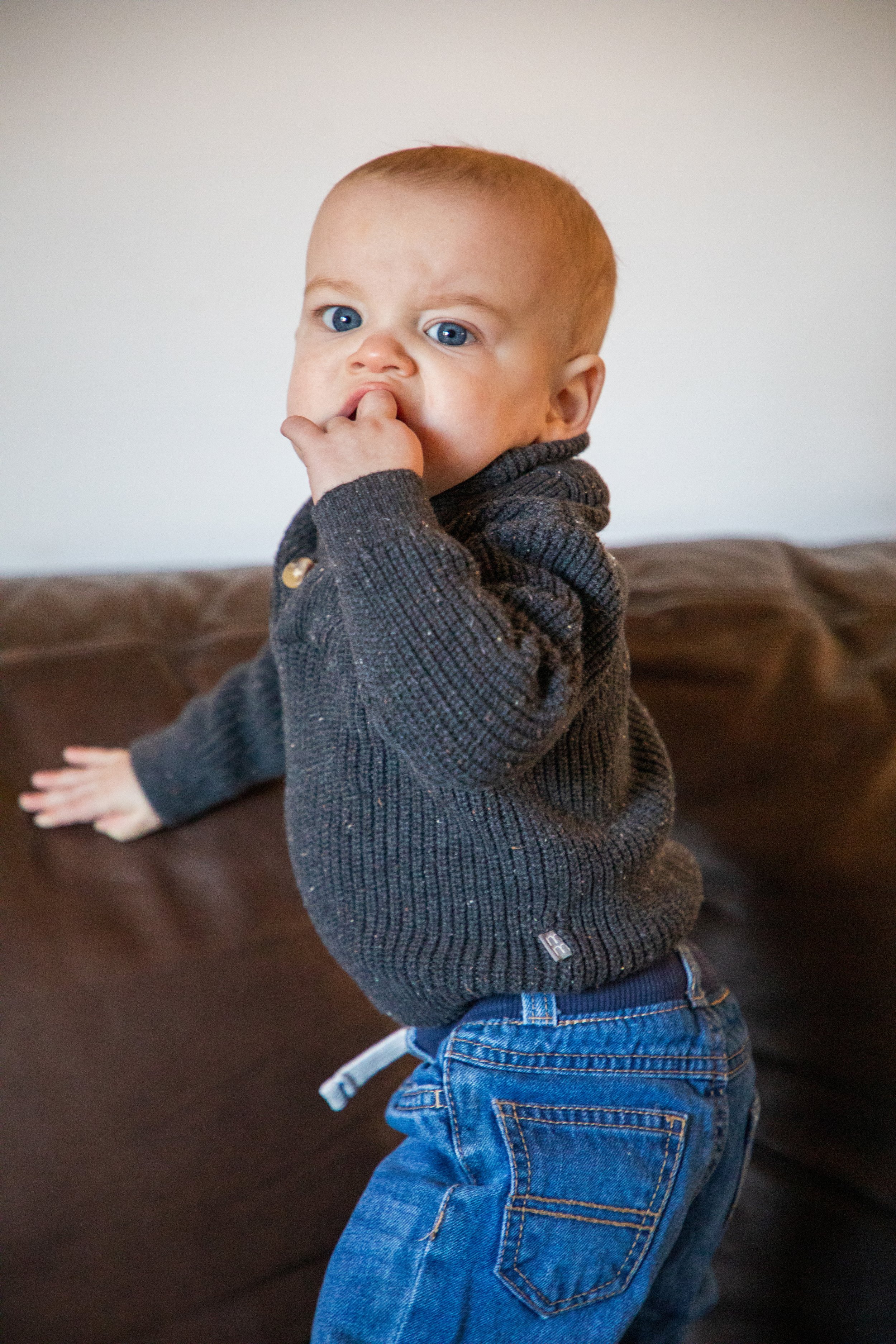 Newborn Baby Photography by Will Locke. A young boy with blue eyes, wearing a dark gray sweater and blue jeans, standing by a brown couch and putting his thumb in his mouth.