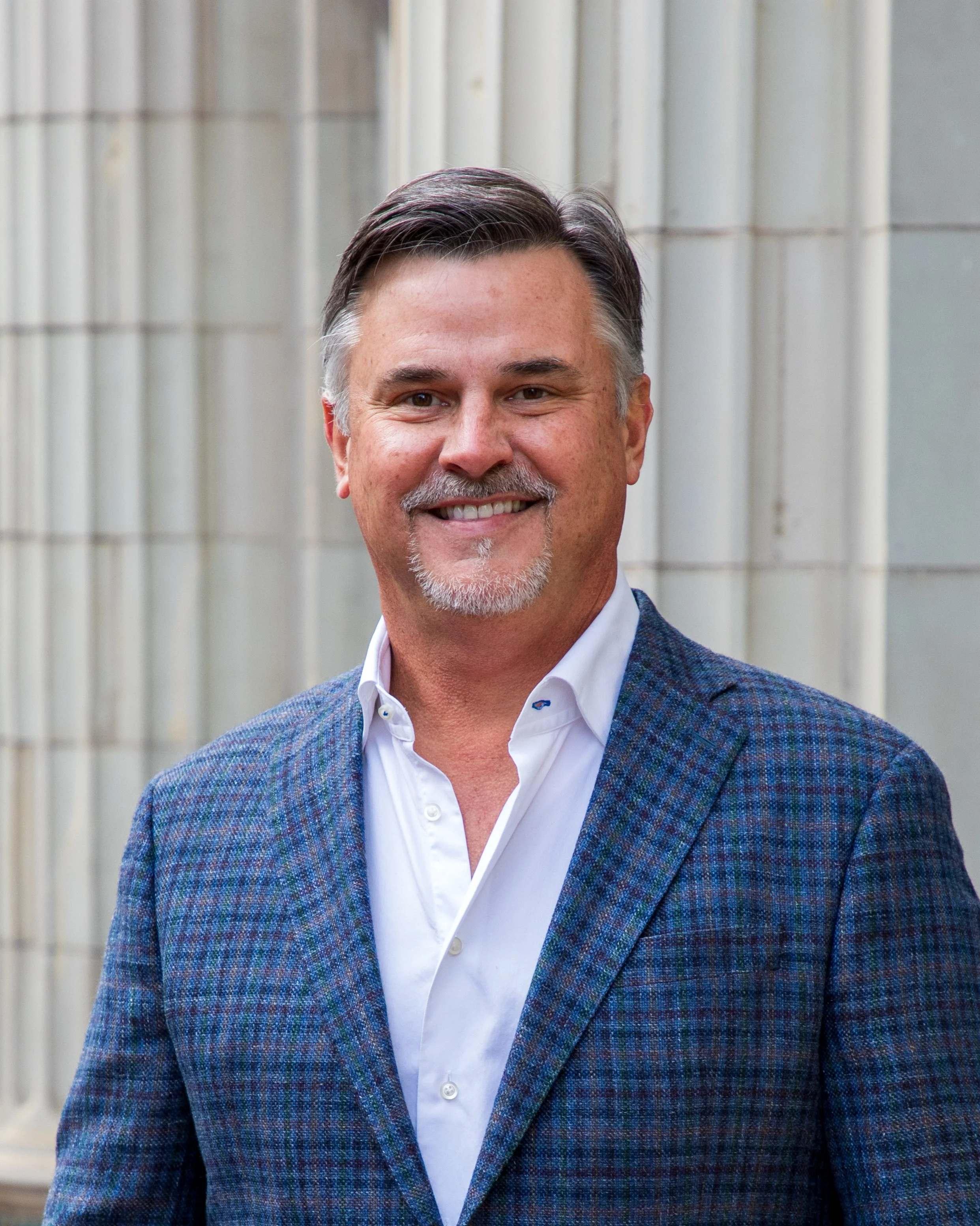 Commercial Headshot Photography by Will Locke. A smiling middle-aged man with gray hair and a goatee, wearing a white dress shirt and a blue plaid blazer, standing outdoors in front of a stone building.