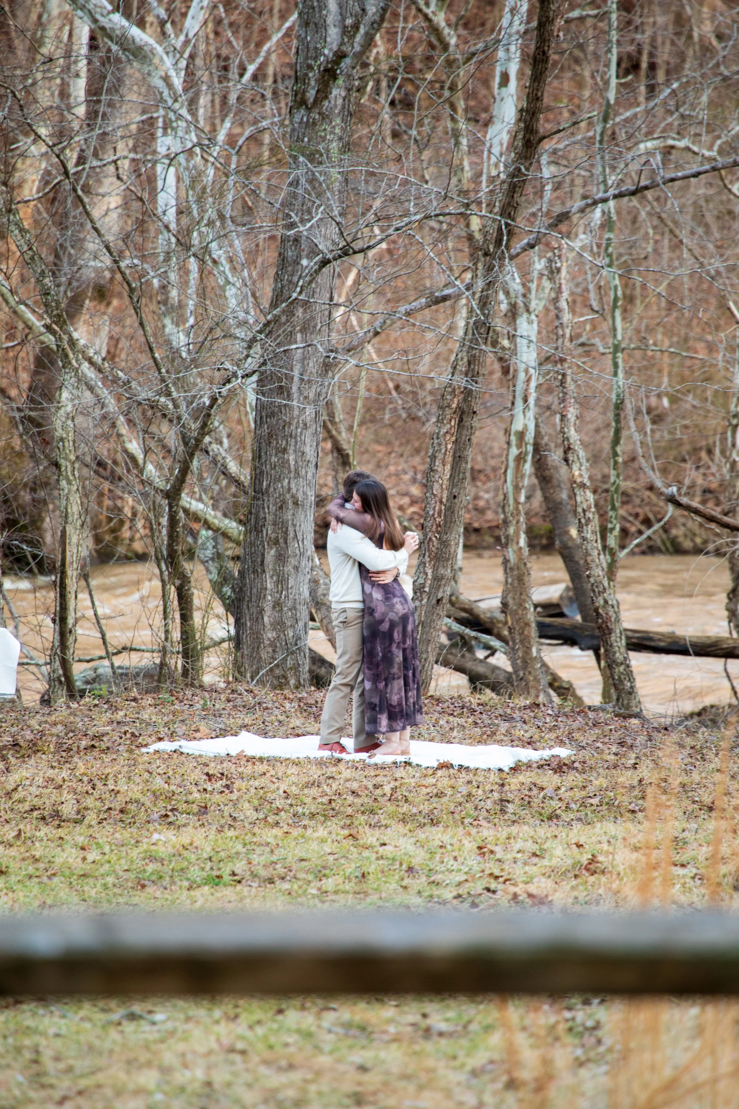 Engagement Photography by Will Locke near Richmond, VA in Montpelier. A couple hugging each other outdoors near a river, standing on a white blanket on the ground, surrounded by leafless trees in a forested area.