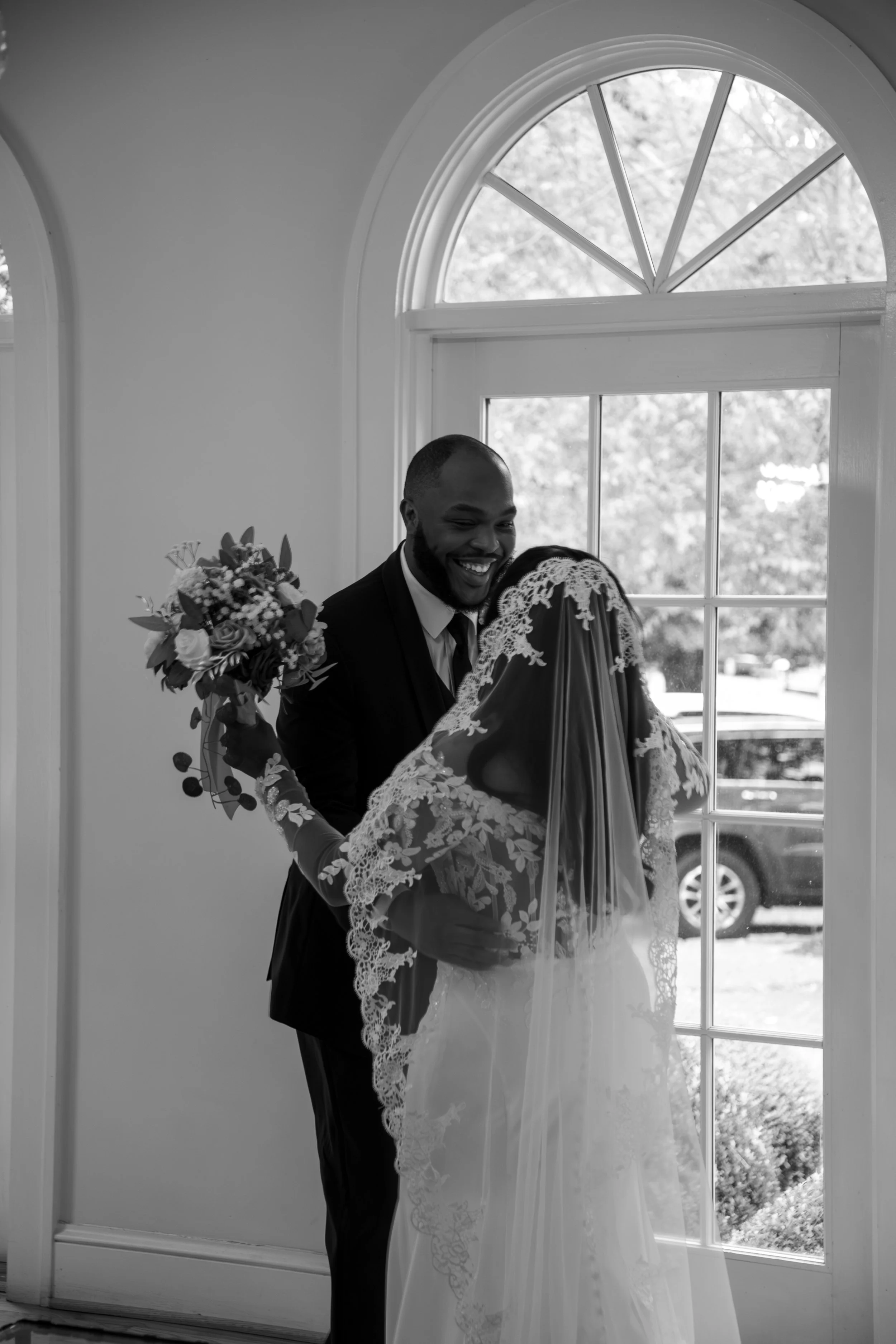 Wedding Photography by Will Locke. A happy bride and groom share a tender moment by a large arched window, with the bride holding a bouquet and both dressed in wedding attire.