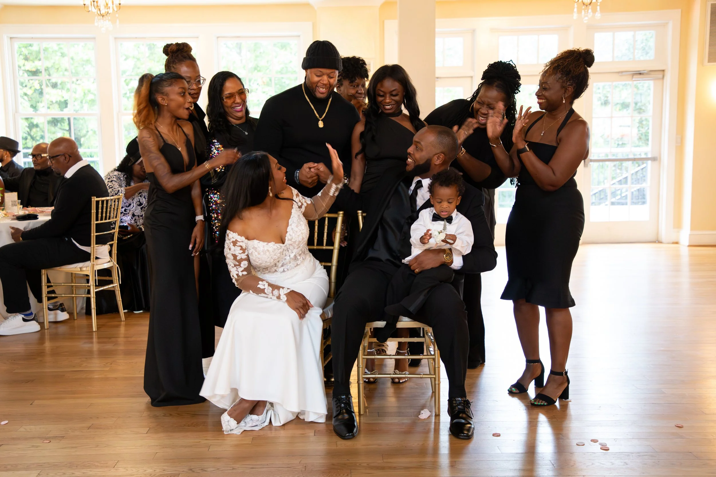 Wedding Photography by Will Locke. A group of people celebrating a wedding, with a bride in a white dress, a groom in a black suit, and a young boy in formal attire. The group is smiling, laughing, and raising their hands in celebration.