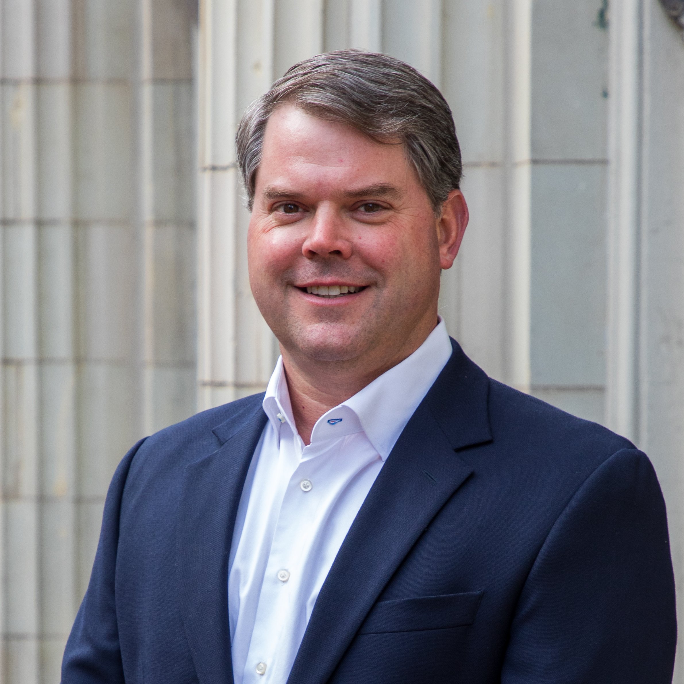 Headshot Photography by Will Locke. A man with short brown hair wearing a navy blazer and white collared shirt stands in front of a stone building, smiling.