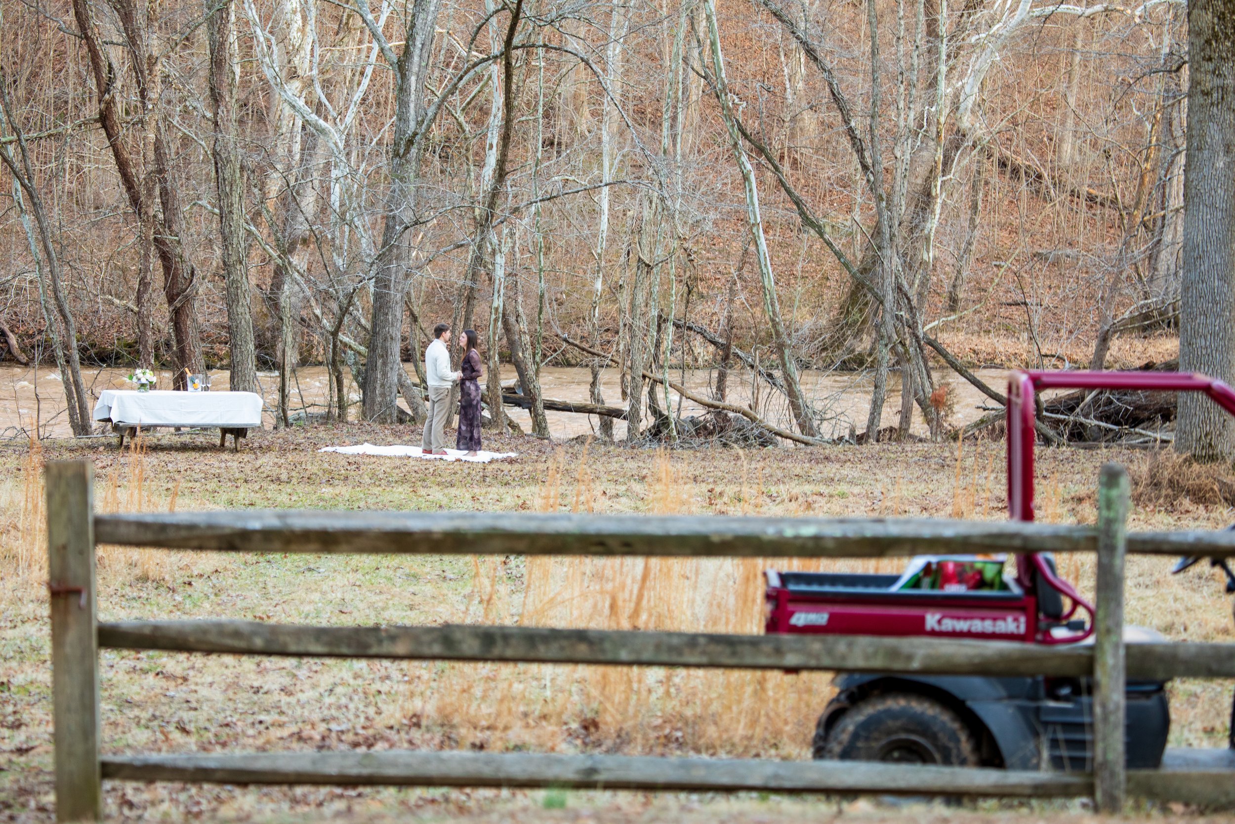 Engagement Photography by Will Locke near Richmond, VA in Montpelier. A couple walks on a white cloth on the ground near a table with tablecloth in a wooded area with a stream on a fall day, with a Kawasaki utility vehicle.