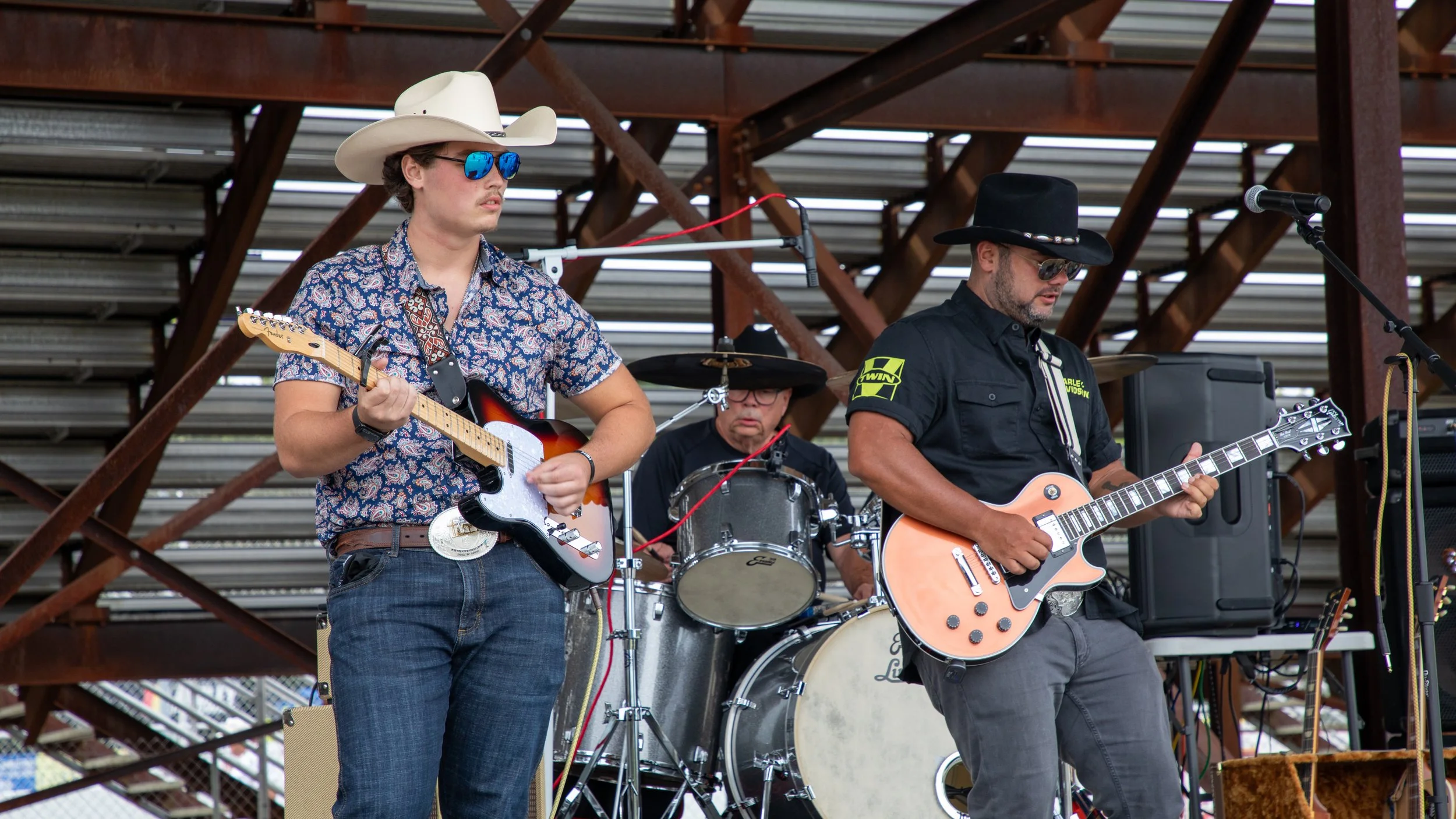 Concert Photography by Will Locke. A band performing outdoors, featuring a young man playing an electric guitar; an older drummer behind him; and another man playing an electric guitar.