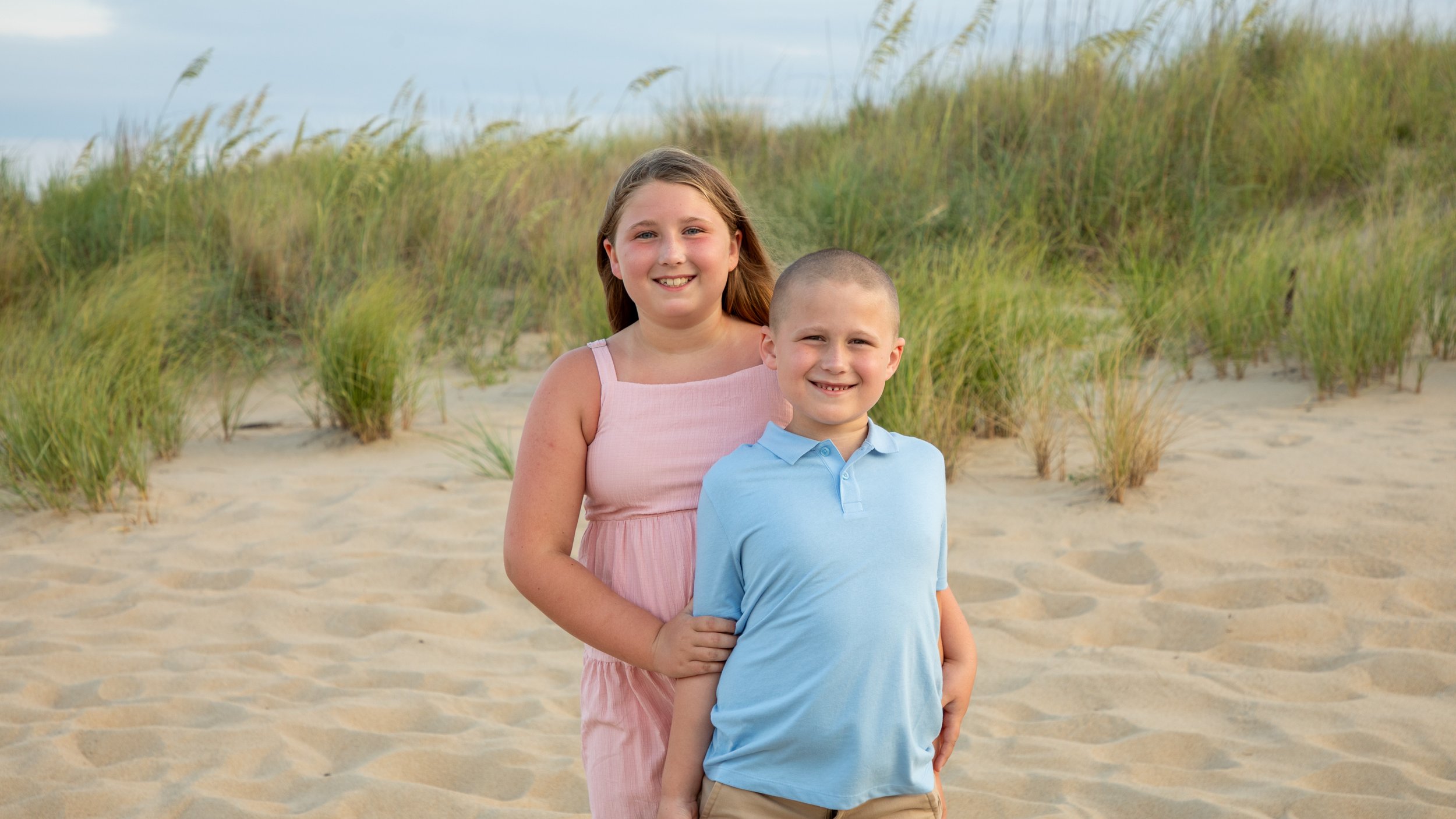 Photo & Video by Will Locke Family Photography in Virginia Beach, VA. Two children, a girl in a pink dress and a boy in a light blue polo shirt, standing on a sandy beach with grassy dunes in the background, smiling at the camera.