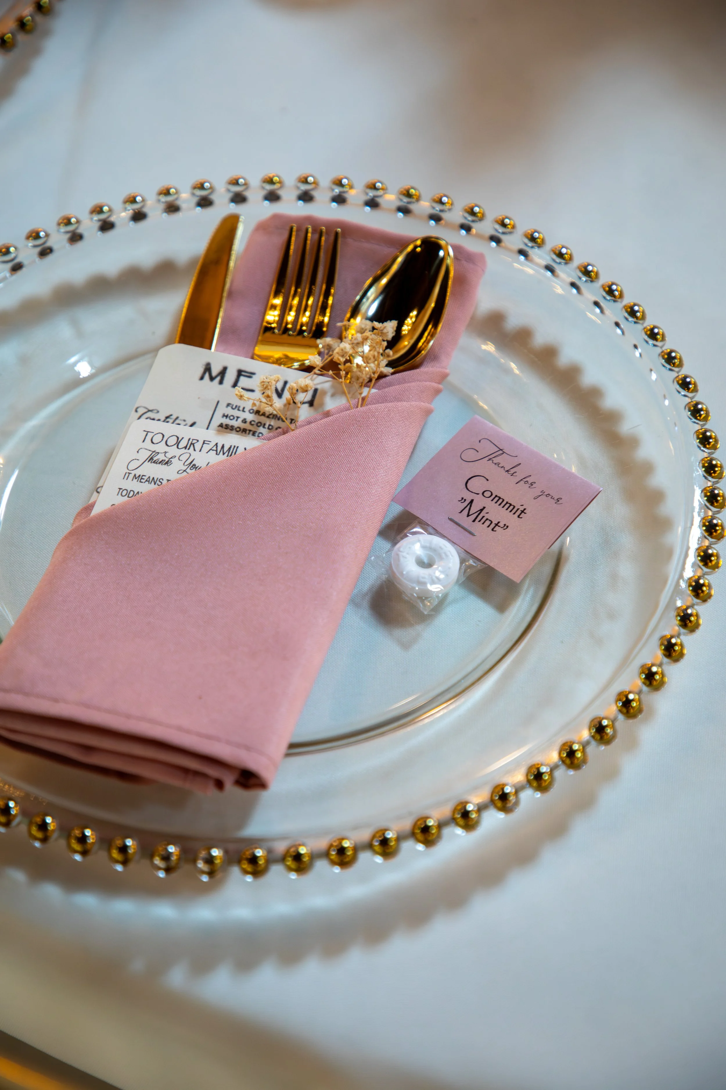 Photo & Video by Will Locke Wedding Photography. Elegant table setting with gold cutlery wrapped in a pink napkin on a clear plate with gold embellishments, a menu, a thank you note, and a small white mints container.