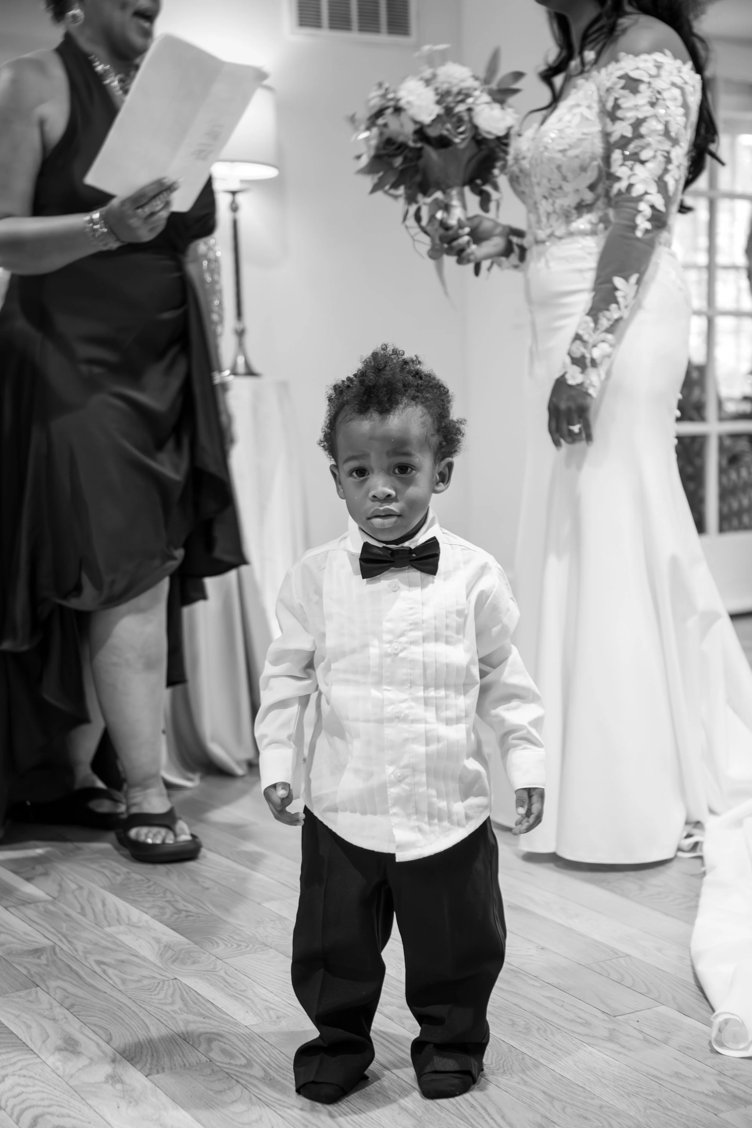 Photo & Video by Will Locke Wedding Photography at the Woman's Club of Portsmouth in VA. A young boy dressed in a white shirt with a bow tie and oversized black pants stands on a wooden floor, looking at the camera. 