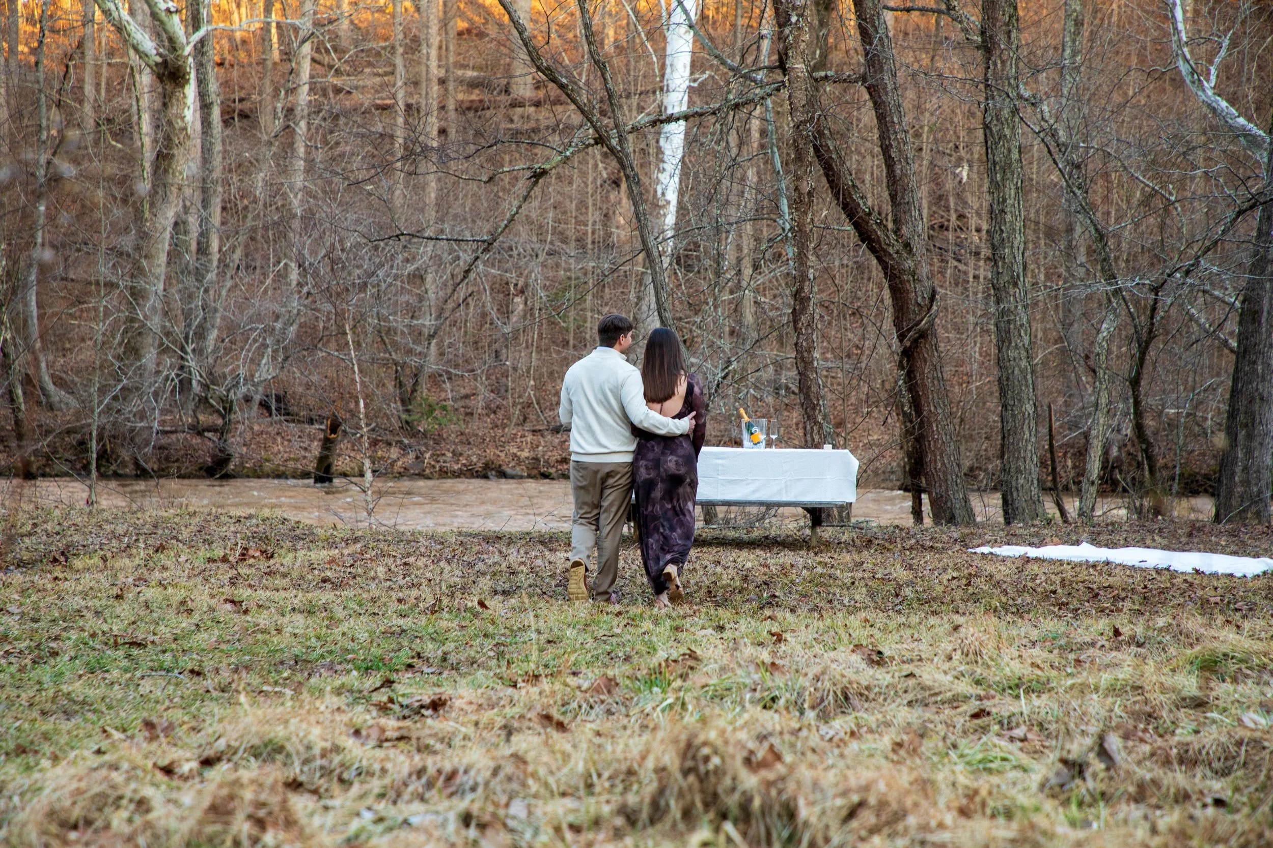 Engagement Photography by Will Locke near Richmond, VA in Montpelier. A couple walks to a white cloth on the ground near a wooded area with a stream on a fall day, with a table on the left side with white tablecloth.