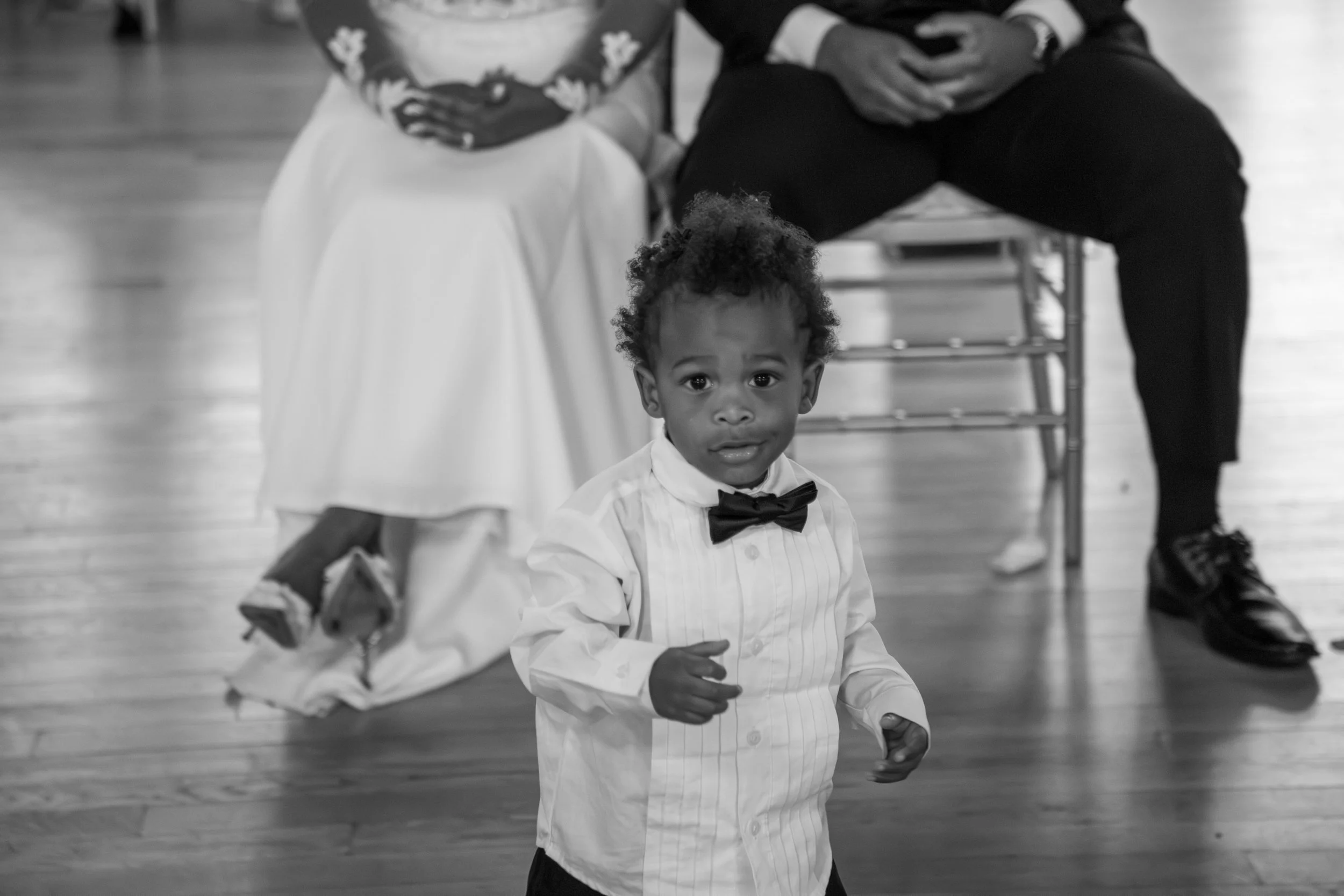Photo & Video by Will Locke Wedding Photography at the Woman's Club of Portsmouth in Virginia. A young child with curly hair wearing a white shirt and a bow tie looking at the camera, with two adults sitting behind him on a wooden floor.