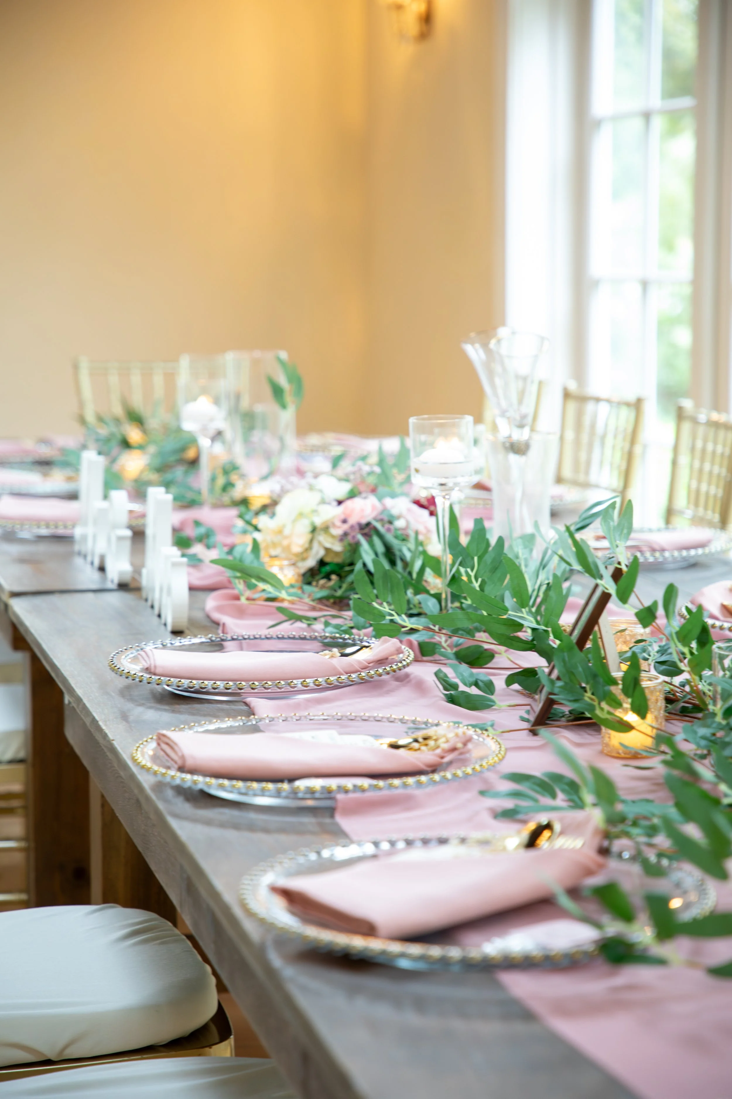Wedding Photography by Will Locke. A decorated dining table with pink napkins, greenery, and gold accents, set for a celebration or event.