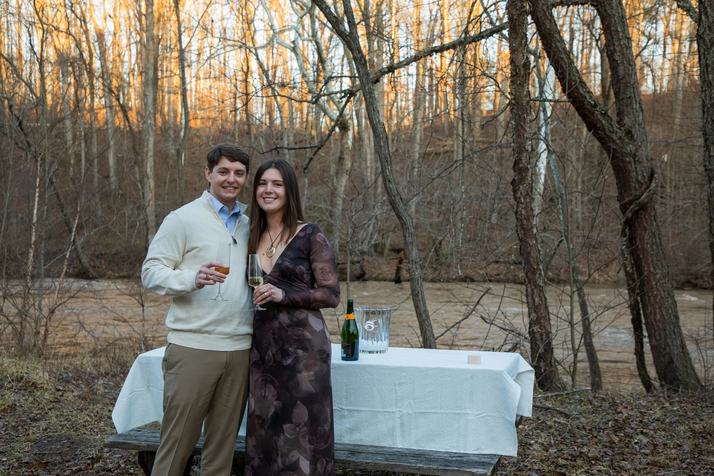 Engagement Photography by Will Locke near Richmond, VA in Montpelier. A man and woman standing together outdoors in front of a table with drinks, smiling at the camera, in a wooded area during sunset.