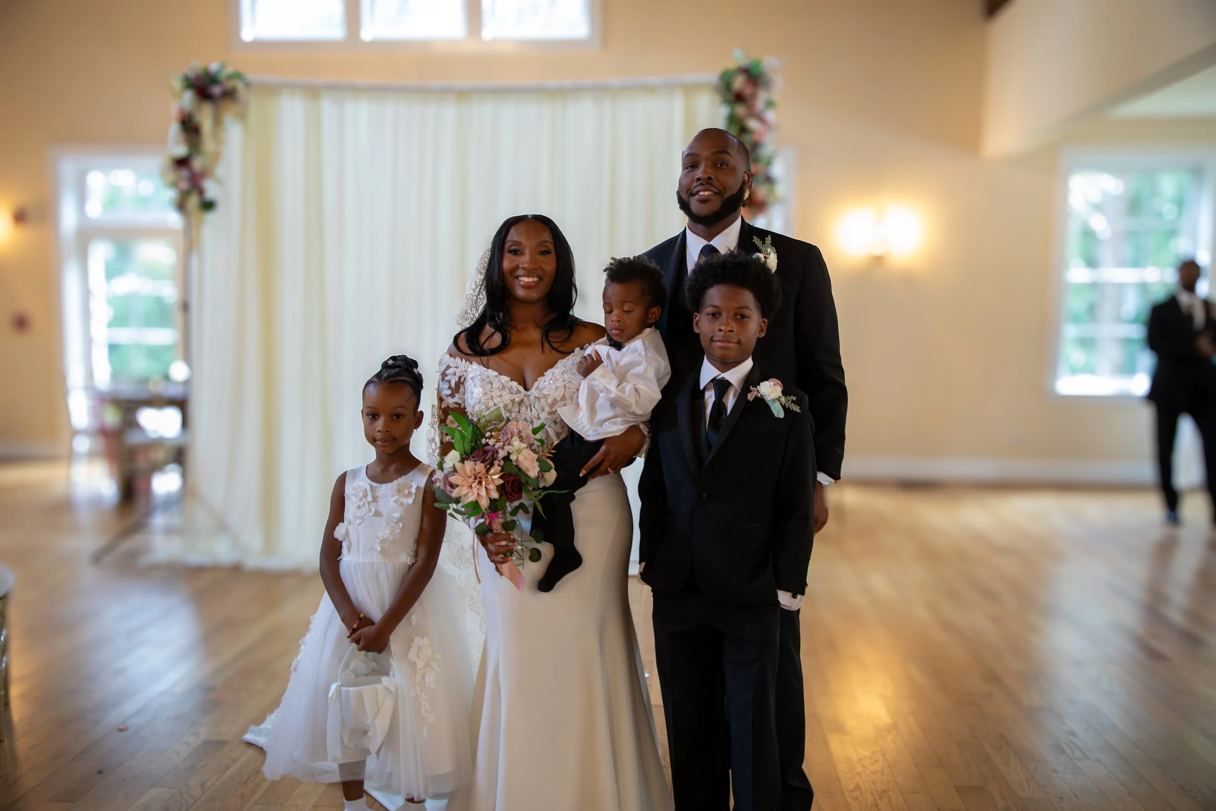 Photo & Video by Will Locke Wedding Photography at the Woman's Club of Portsmouth. A family dressed in wedding attire standing together in a decorated reception hall, with a bride and groom at the center surrounded by children and a floral backdrop.