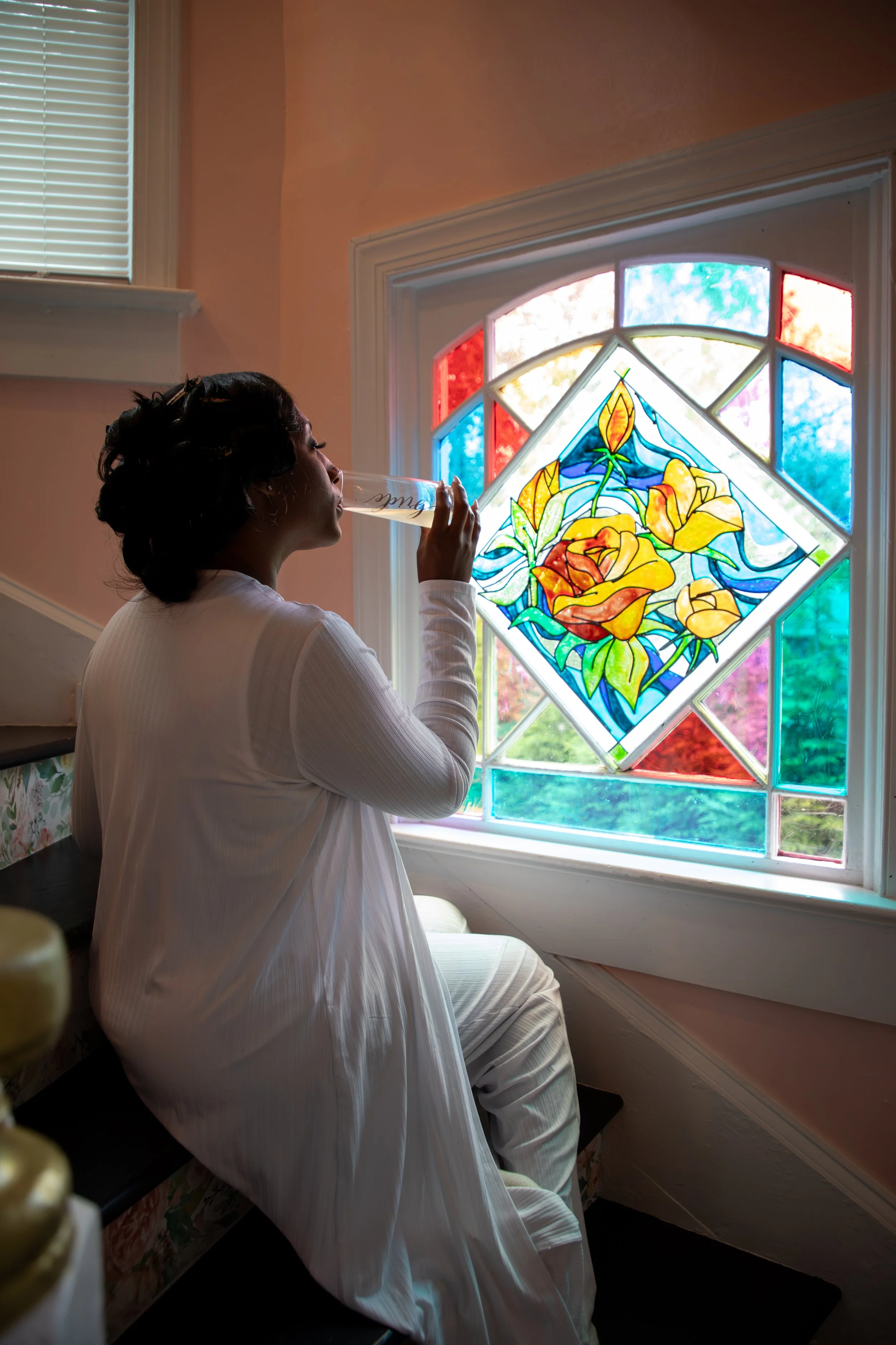 Photo & Video by Will Locke Wedding Photography at the Bridgerton House in Portsmouth, VA. A woman dressed in white sitting on stairs, drinking from a clear cup, next to a colorful stained glass window with floral design.