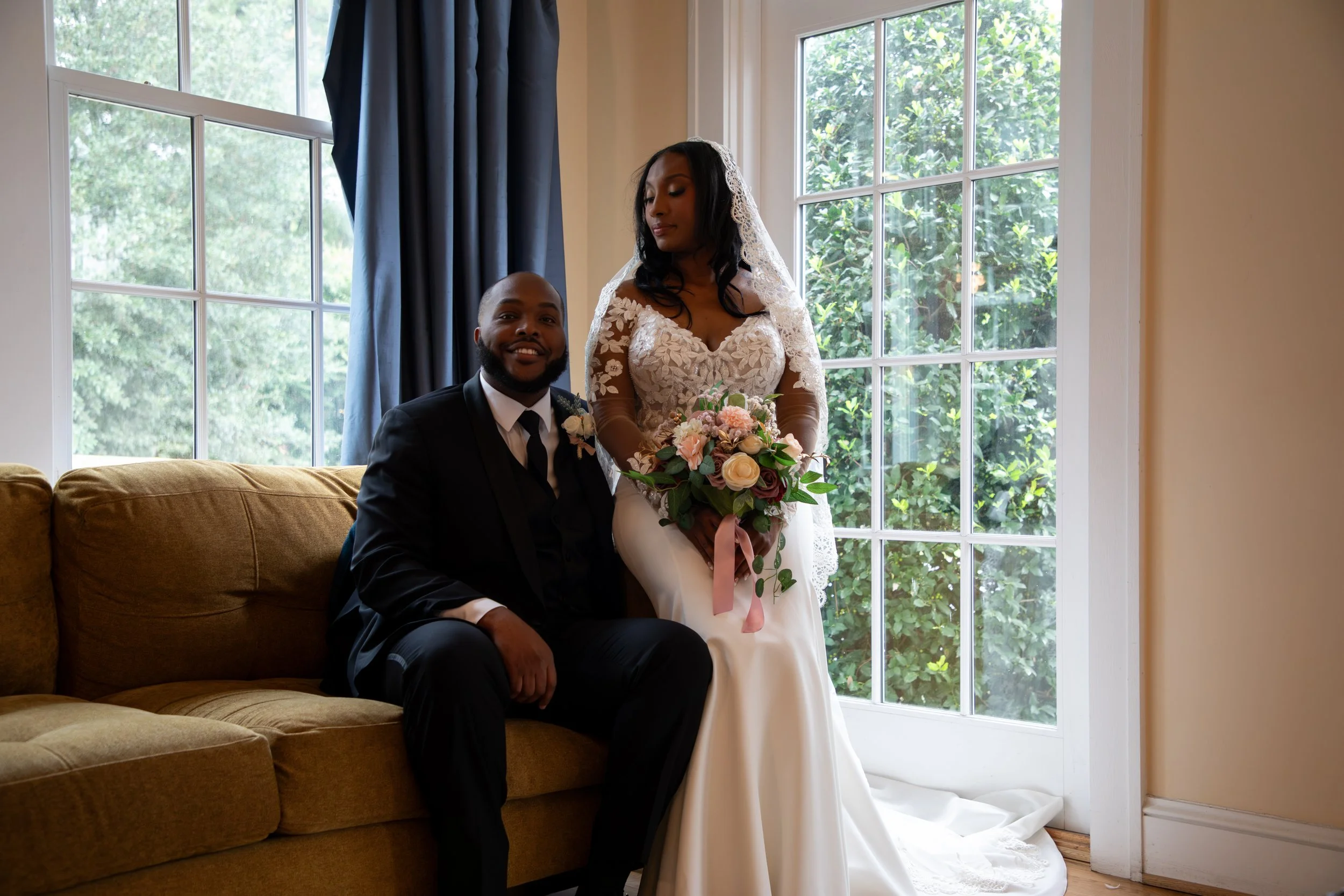 Photo & Video by Will Locke Wedding Photography at the Woman's Club of Portsmouth in VA. A newlywed couple sitting near a large window with lush greenery outside. The bride is standing, holding a bouquet, wearing a lace wedding dress with a veil. 