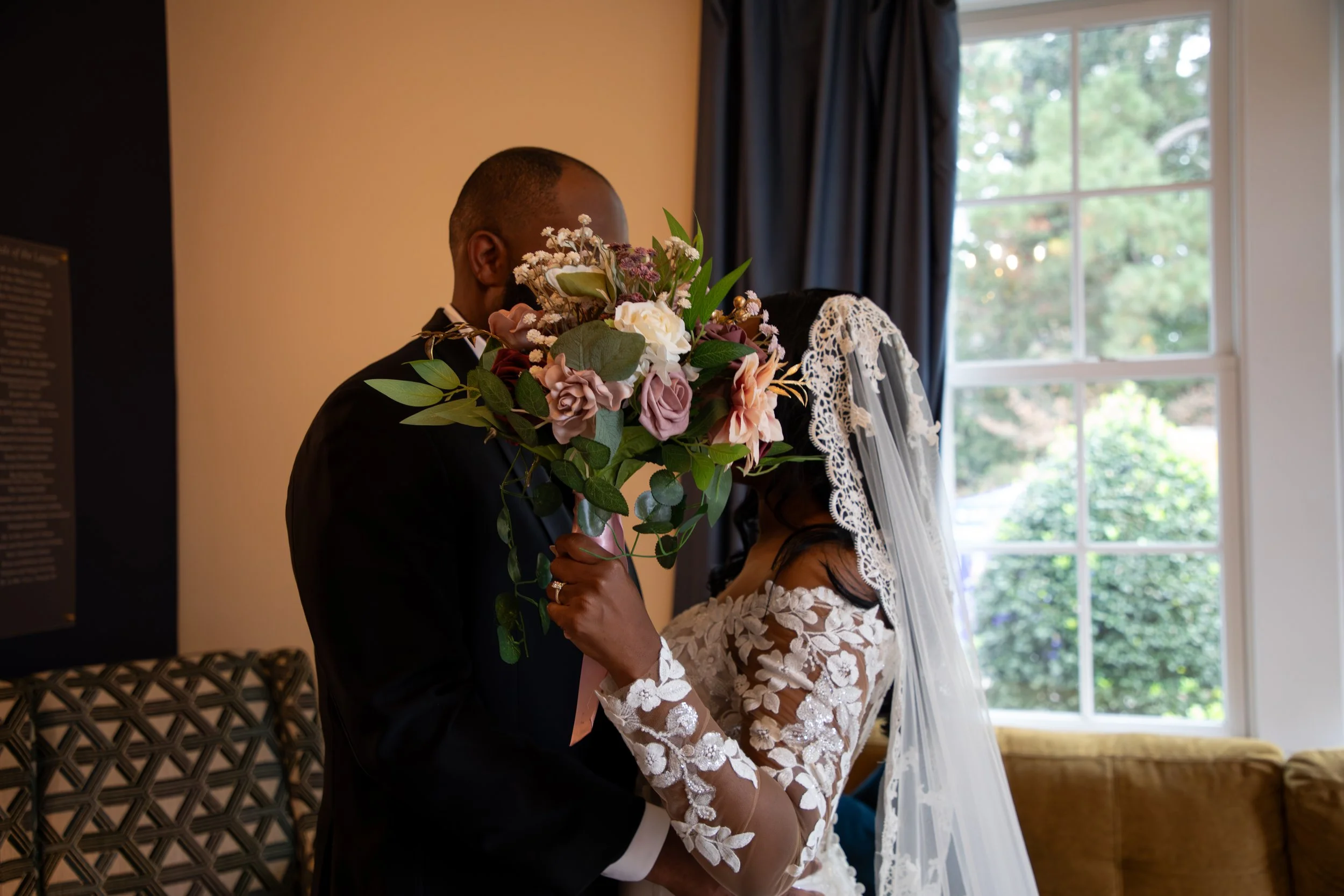 Wedding Photography by Will Locke. A bride and groom in wedding attire sharing a kiss behind a bouquet of flowers, indoors near a window.