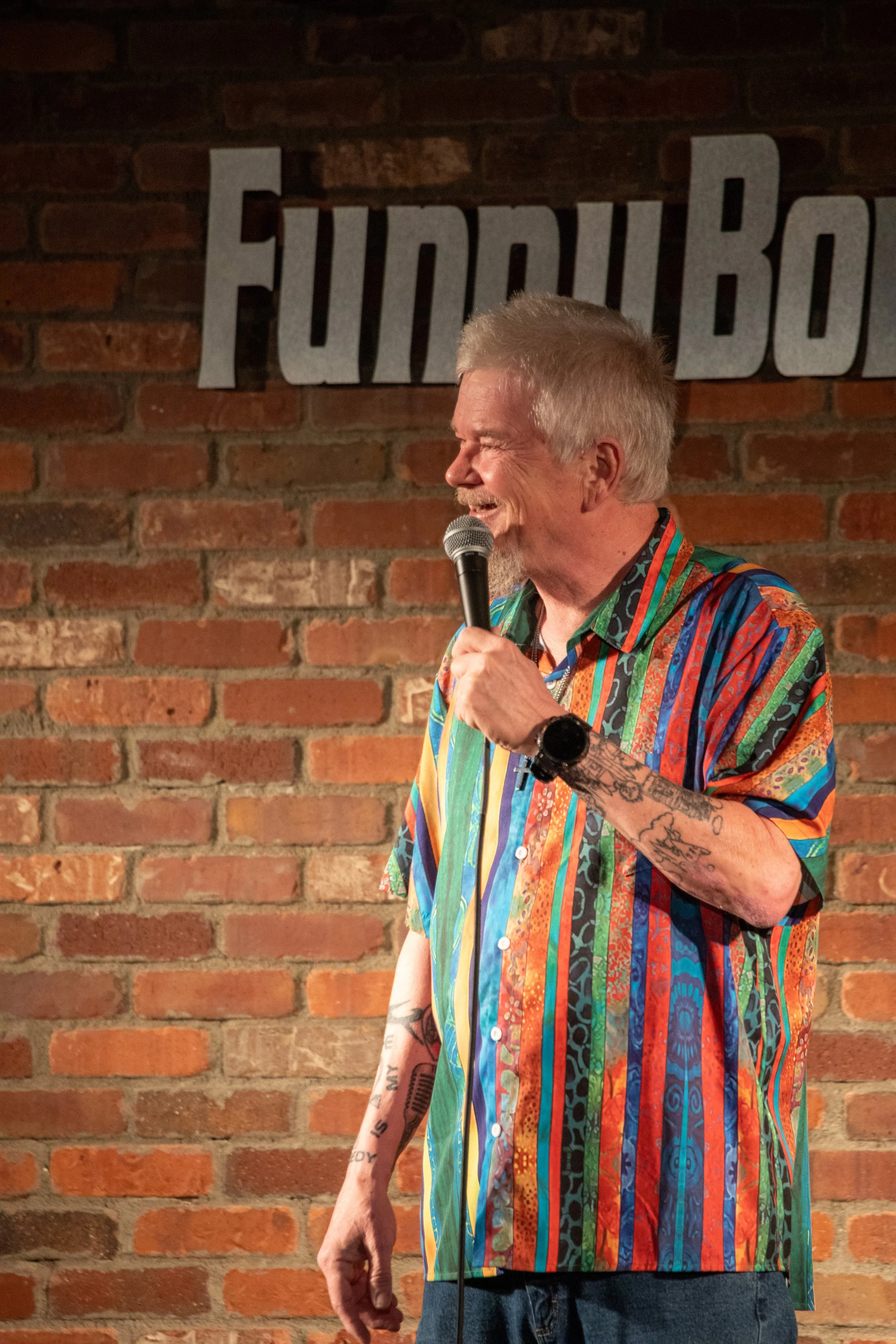 Comedy Photography by Will Locke. A man with tattoos, wearing a colorful, striped shirt, speaking into a microphone on stage at a comedy club with a brick wall background and a sign that says "FUNNY BONE."