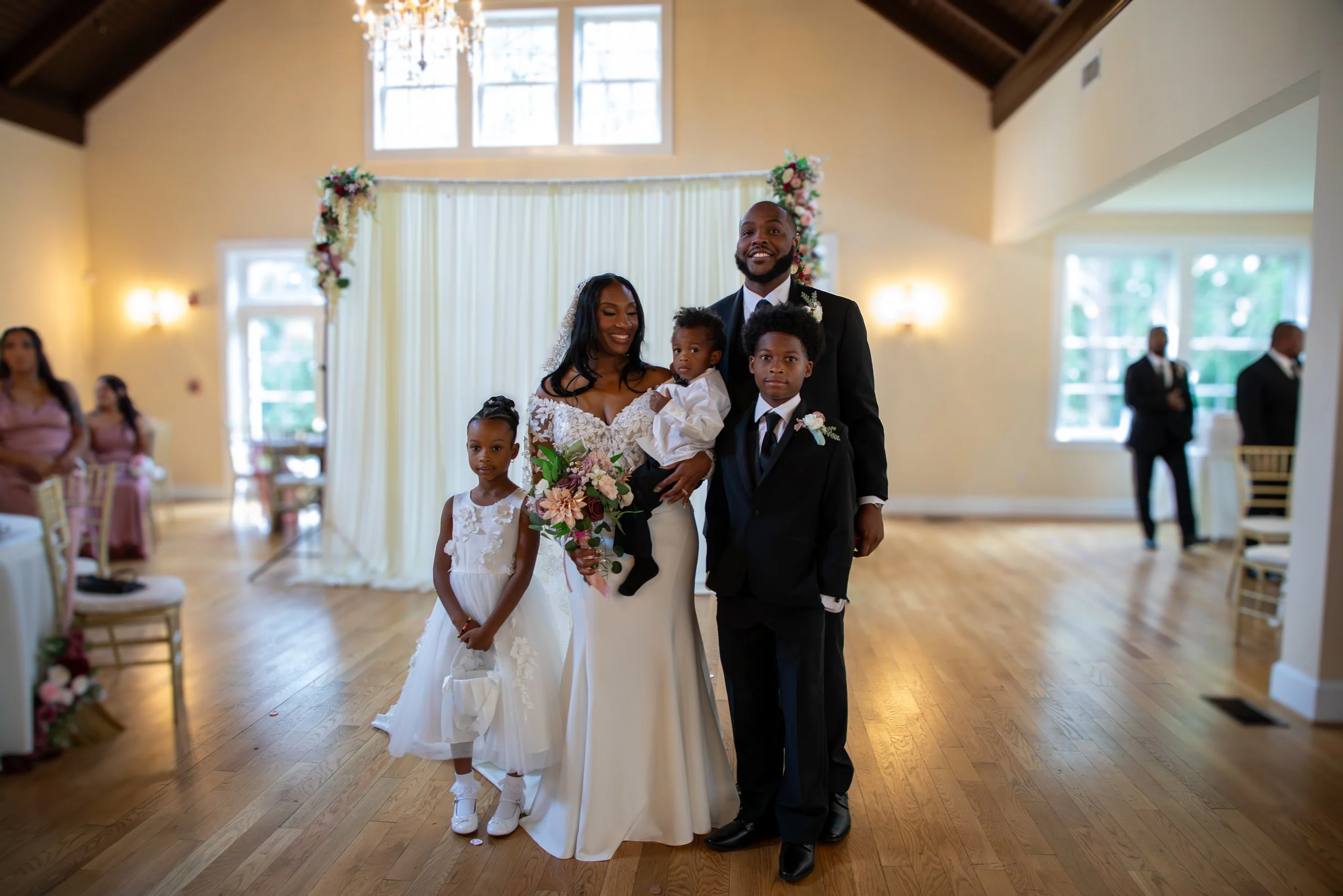 A newlywed couple with three children standing in a decorated wedding venue. Photo & Video by Will Locke Wedding Photography at the Woman's Club of Portsmouth in Virginia. 