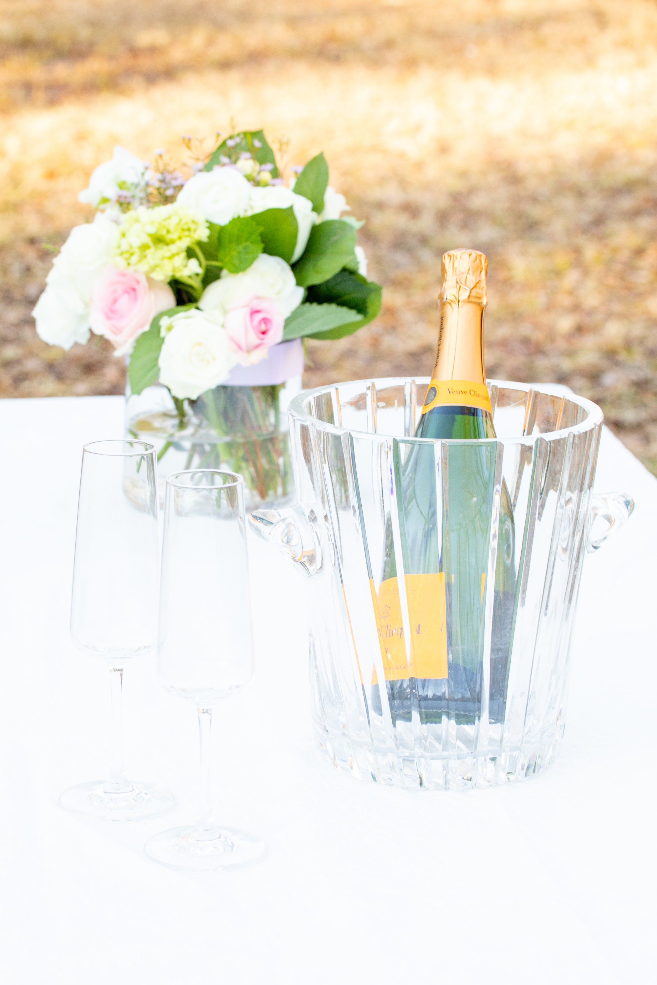 Engagement Photography by Will Locke near Richmond, VA in Montpelier. A bottle of Veuve Clicquot champagne in an ice bucket with two empty champagne flutes on a white table outdoors, with a flower arrangement. 