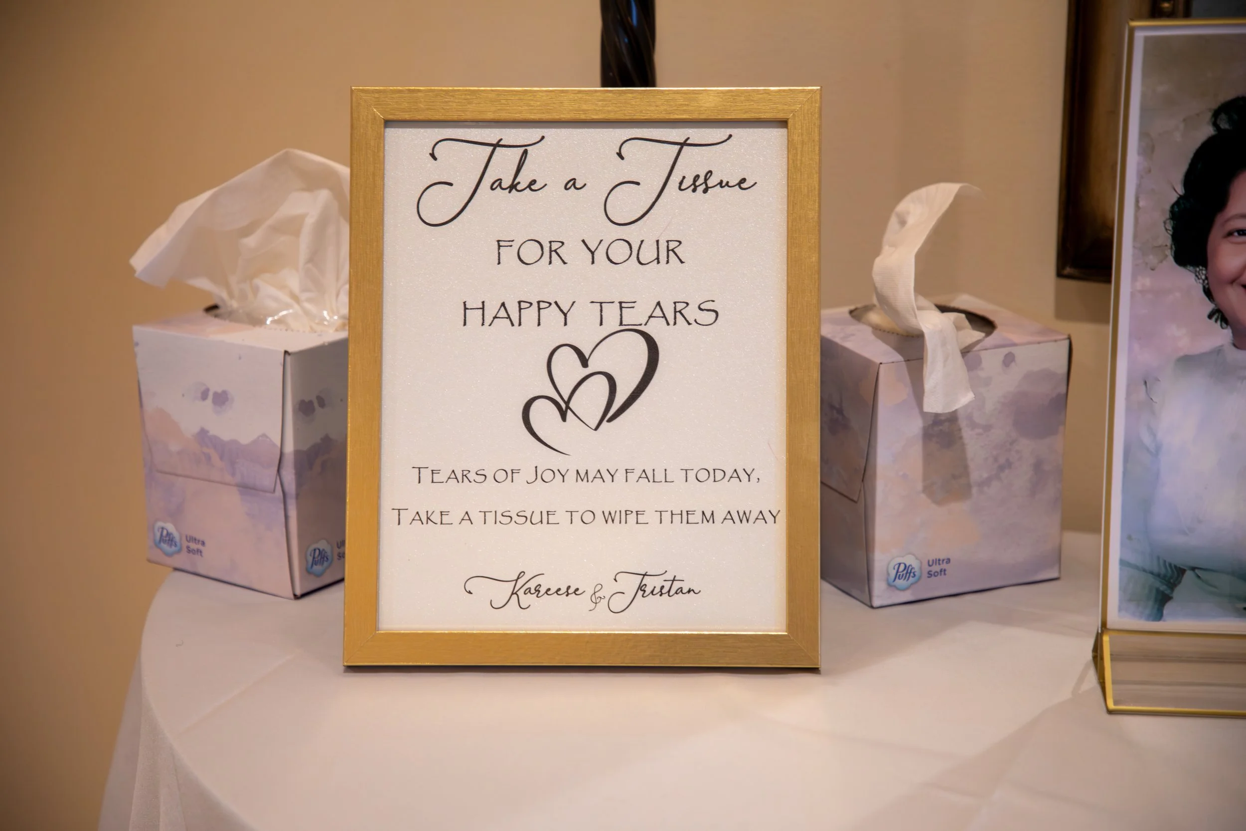 Sign with a golden frame on a table reading: "Take a Tissue for your happy tears. Tears of joy may fall today, take a tissue to wipe them away. Kareem & Tristan." Wedding Photography by Will Locke. 