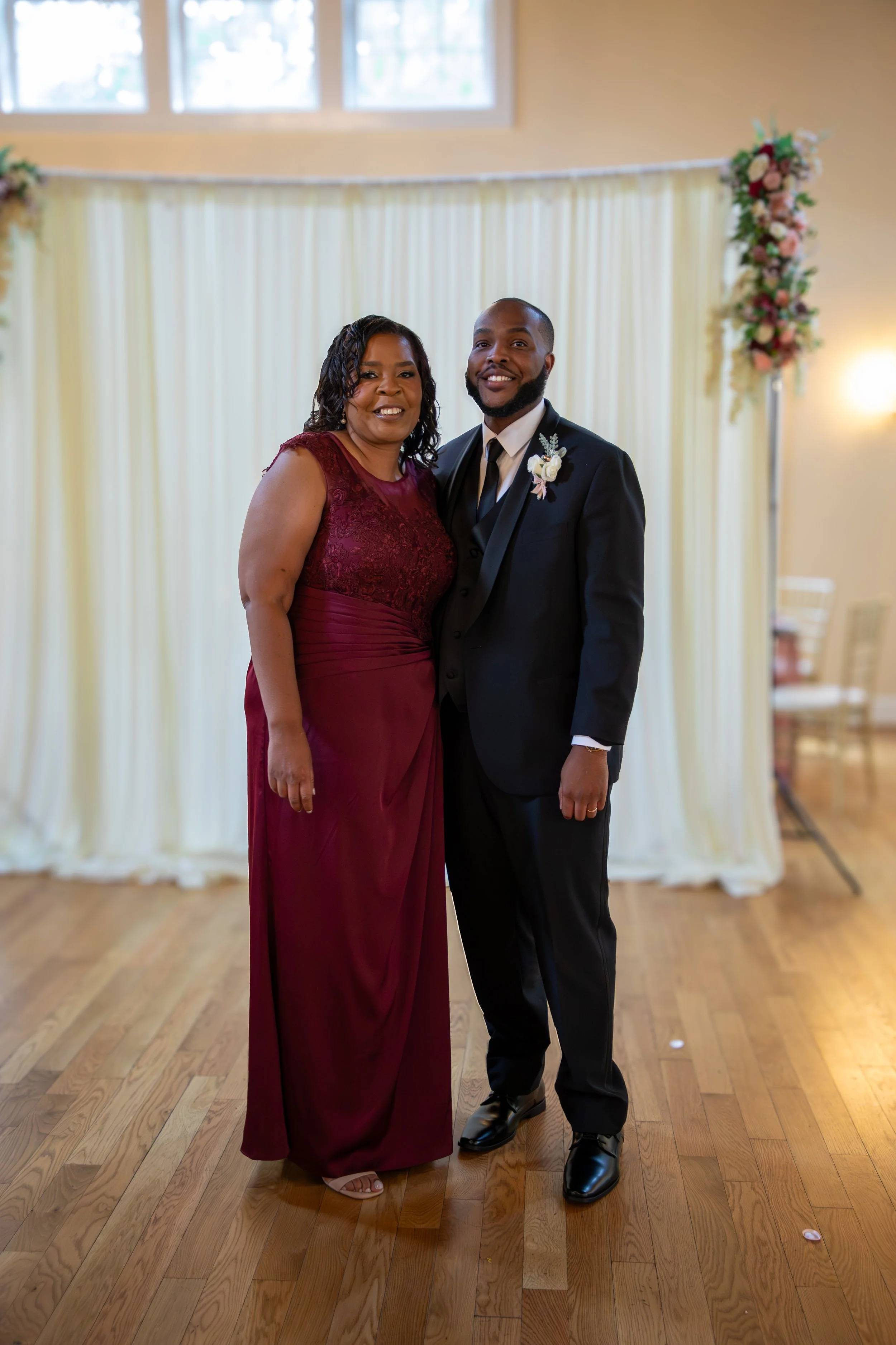 Photo & Video by Will Locke Wedding Photography at the Woman's Club of Portsmouth in VA. A woman in a burgundy dress and a man in a black suit and tie standing together in a decorated indoor venue with wooden flooring and floral arrangements.