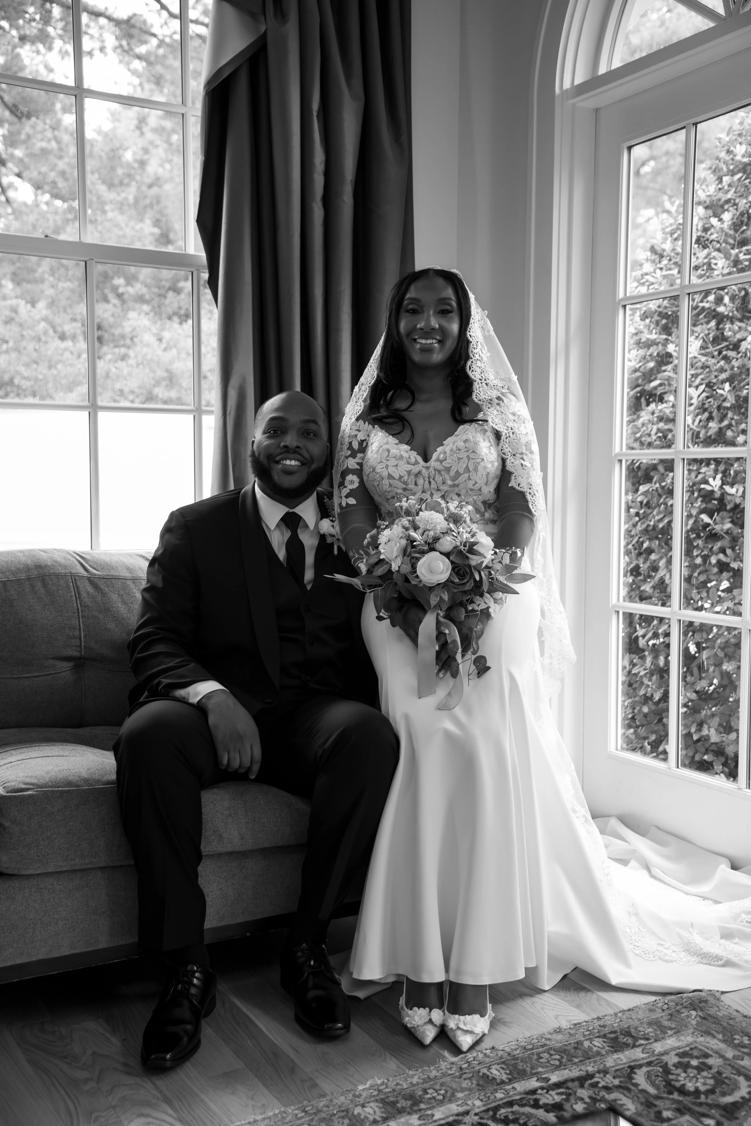 Photo & Video by Will Locke Wedding Photography at the Woman's Club of Portsmouth in Virginia. A bride and groom celebrating their wedding indoors near large windows, with the bride holding a bouquet and wearing a lace wedding gown and veil.