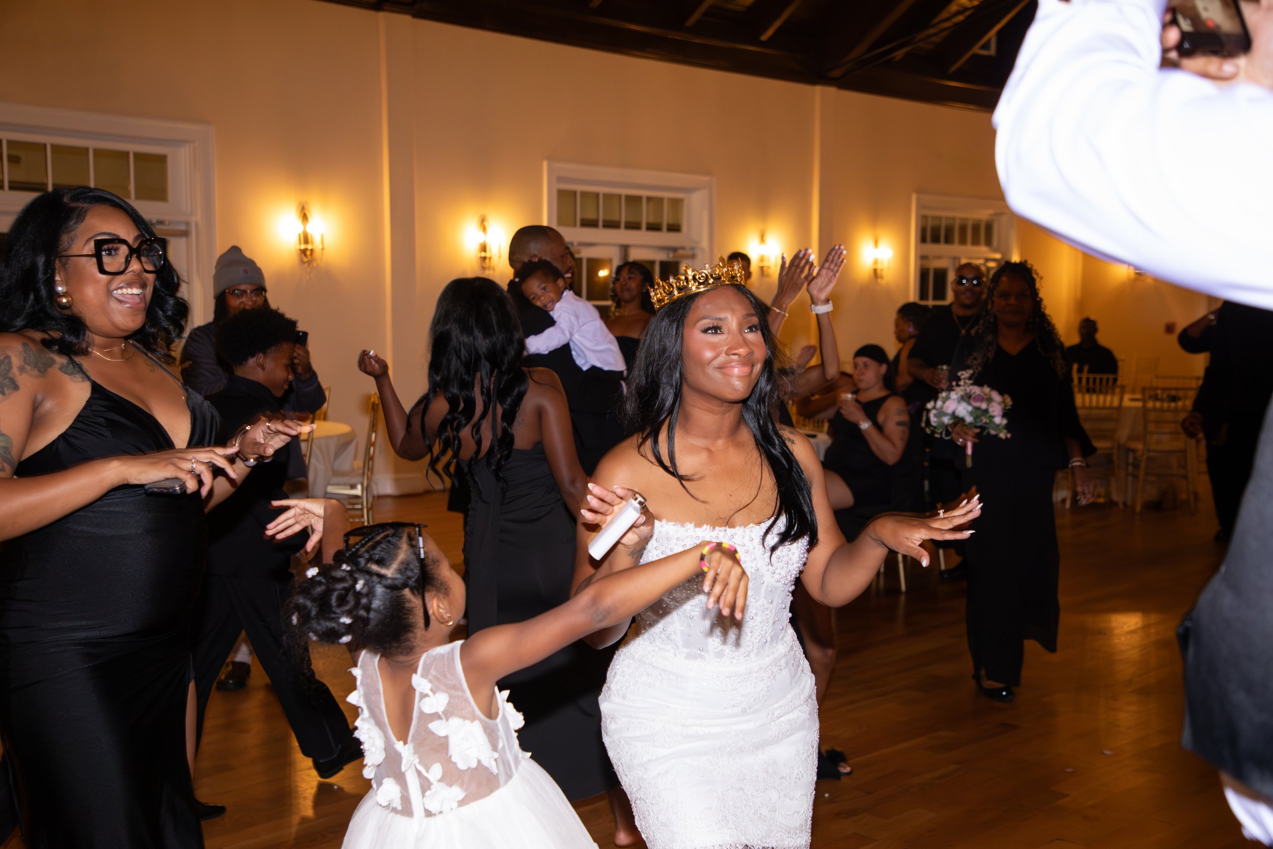 Wedding Photography by Will Locke. Women and children dancing and having fun at a wedding reception in a decorated hall, with warm lighting and elegant decor.