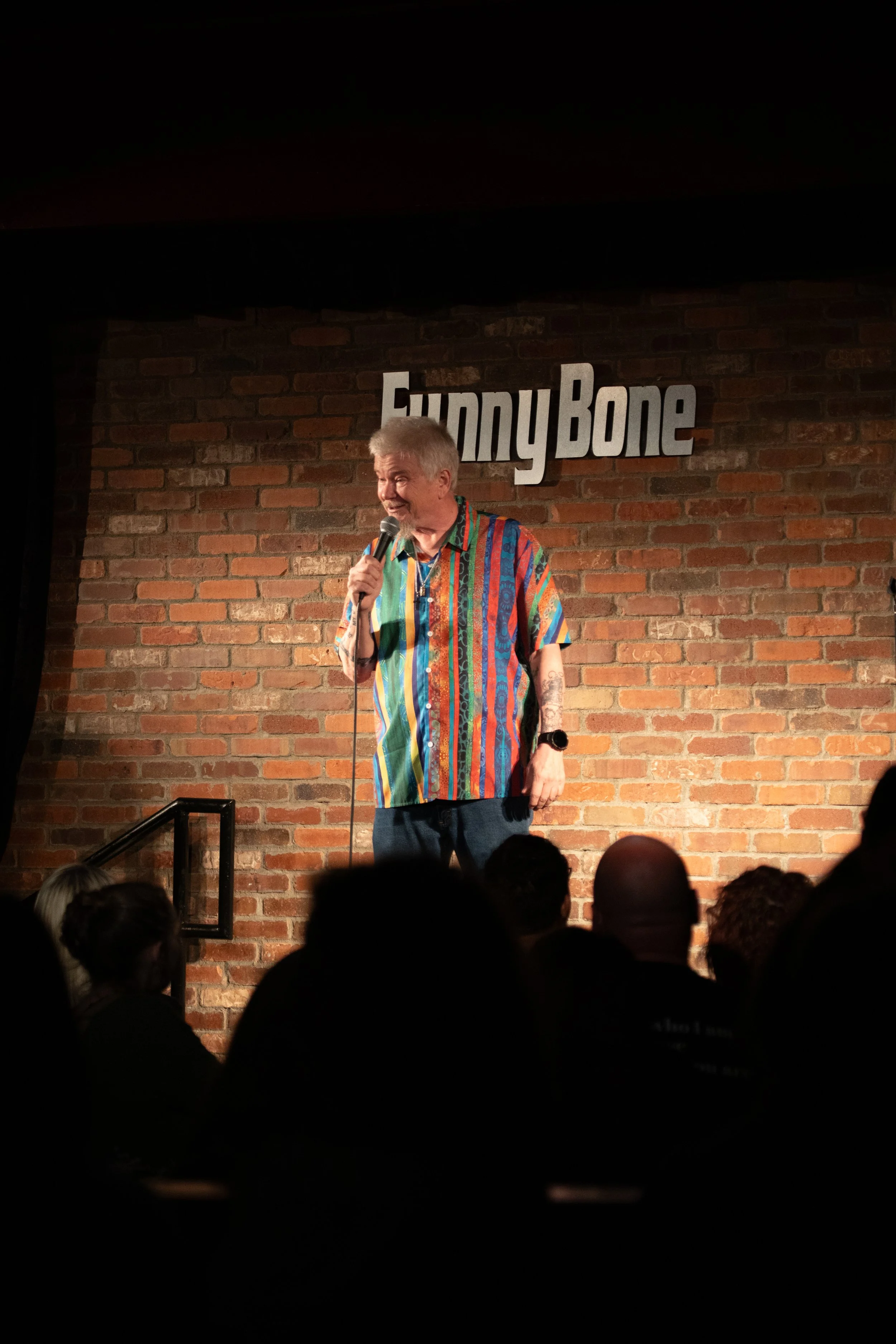 Comedy Photography by Will Locke. A comedian performing on stage at the Sunny Bone comedy club, standing in front of a brick wall with the club's sign, holding a microphone, wearing a colorful striped shirt.