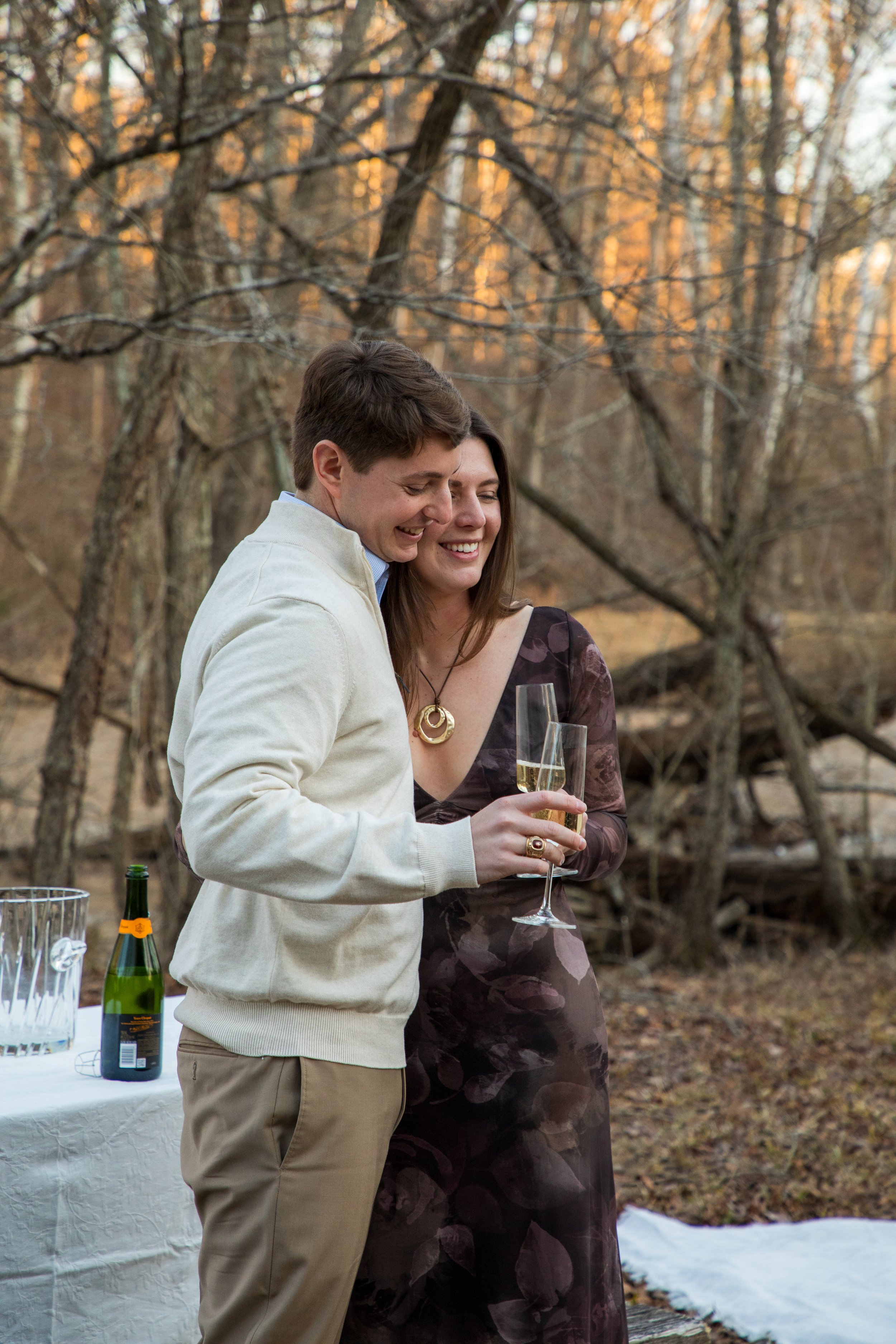 Engagement Photography by Will Locke near Richmond, VA in Montpelier. A man and woman smiling and hugging while holding champagne glasses outdoors during sunset, with a table of flowers nearby.