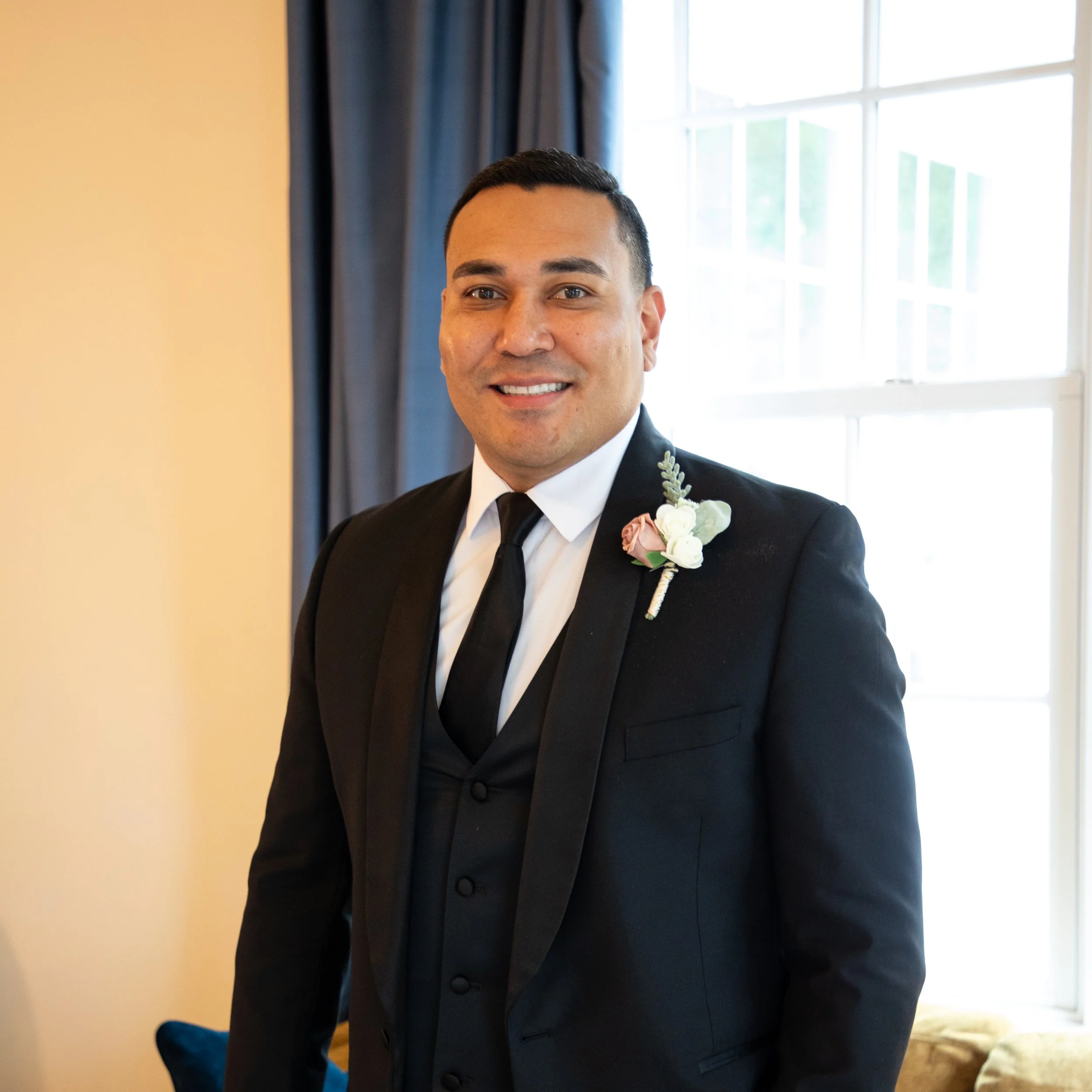 Headshot Photography by Will Locke. A man in a black tuxedo with a white shirt and black tie, smiling, standing indoors near a window with blue curtains, with a boutonniere on his lapel.