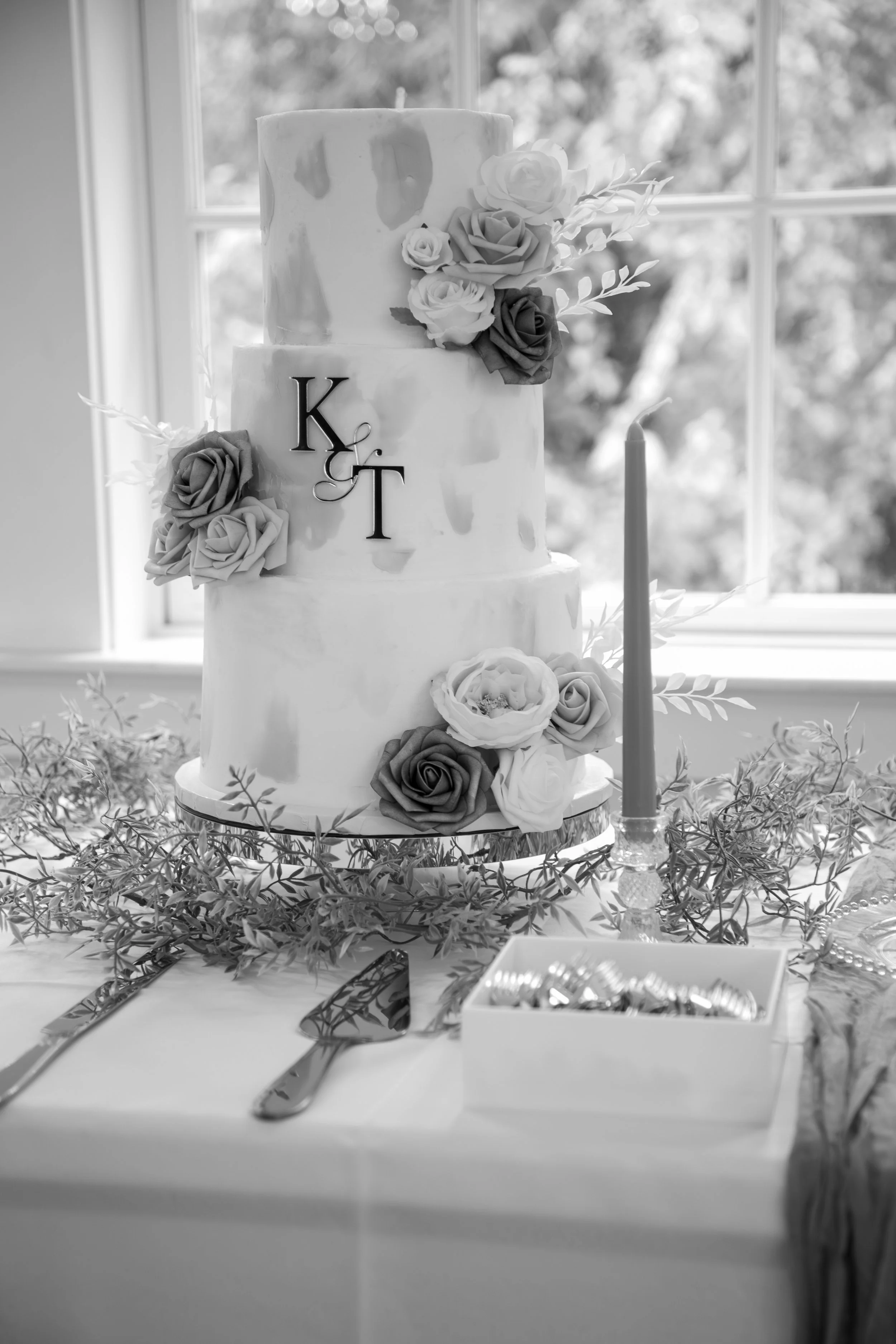Wedding Photography by Will Locke. A three-tier wedding cake decorated with roses and monogram initials 'K & T', displayed on a table with cutlery, a candlestick, and greenery, in front of a window.