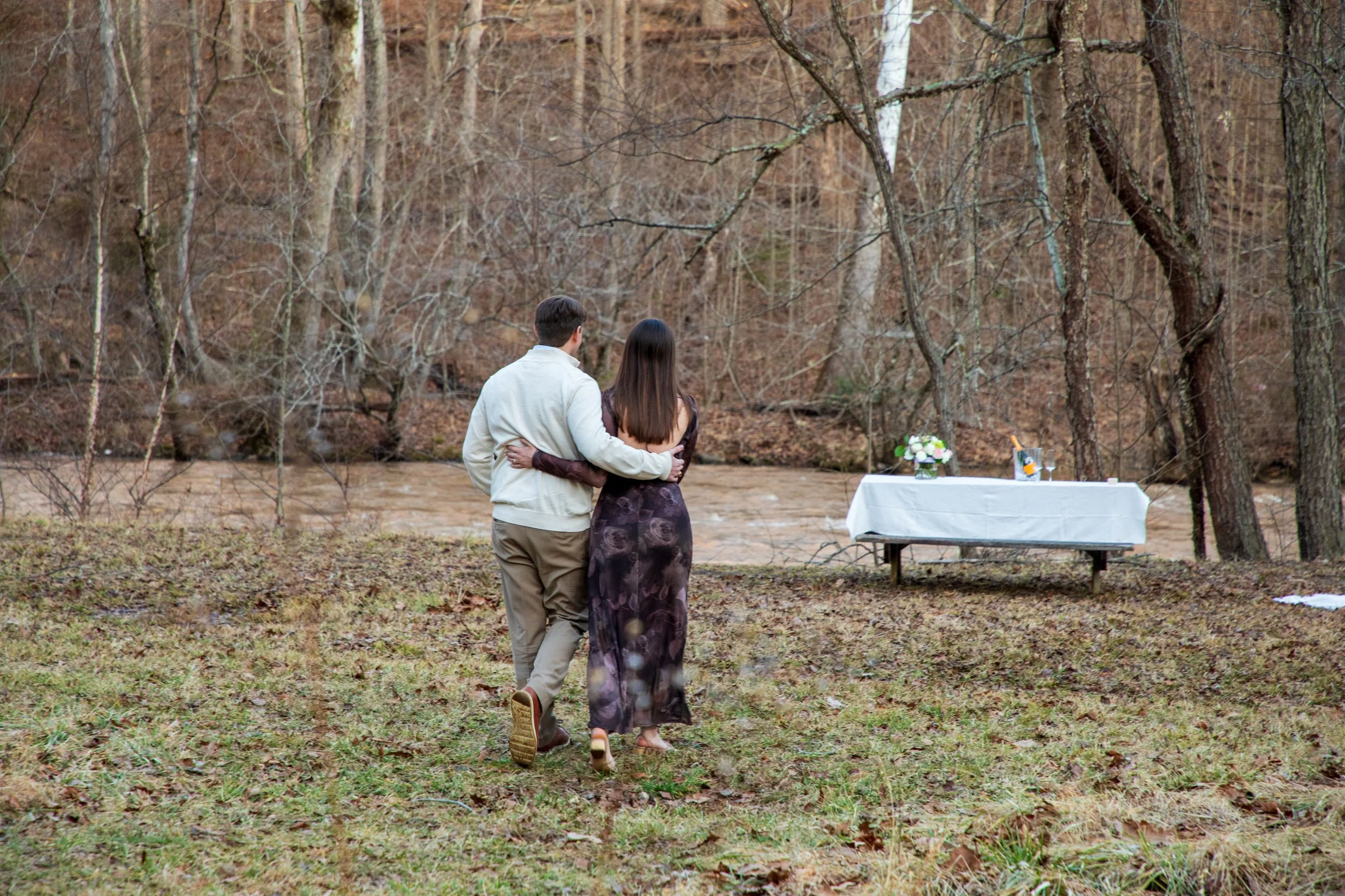 Engagement Photography by Will Locke near Richmond, VA in Montpelier. A couple walks on a white cloth on the ground near a wooded area with a stream on a fall day, with a table on the left side.