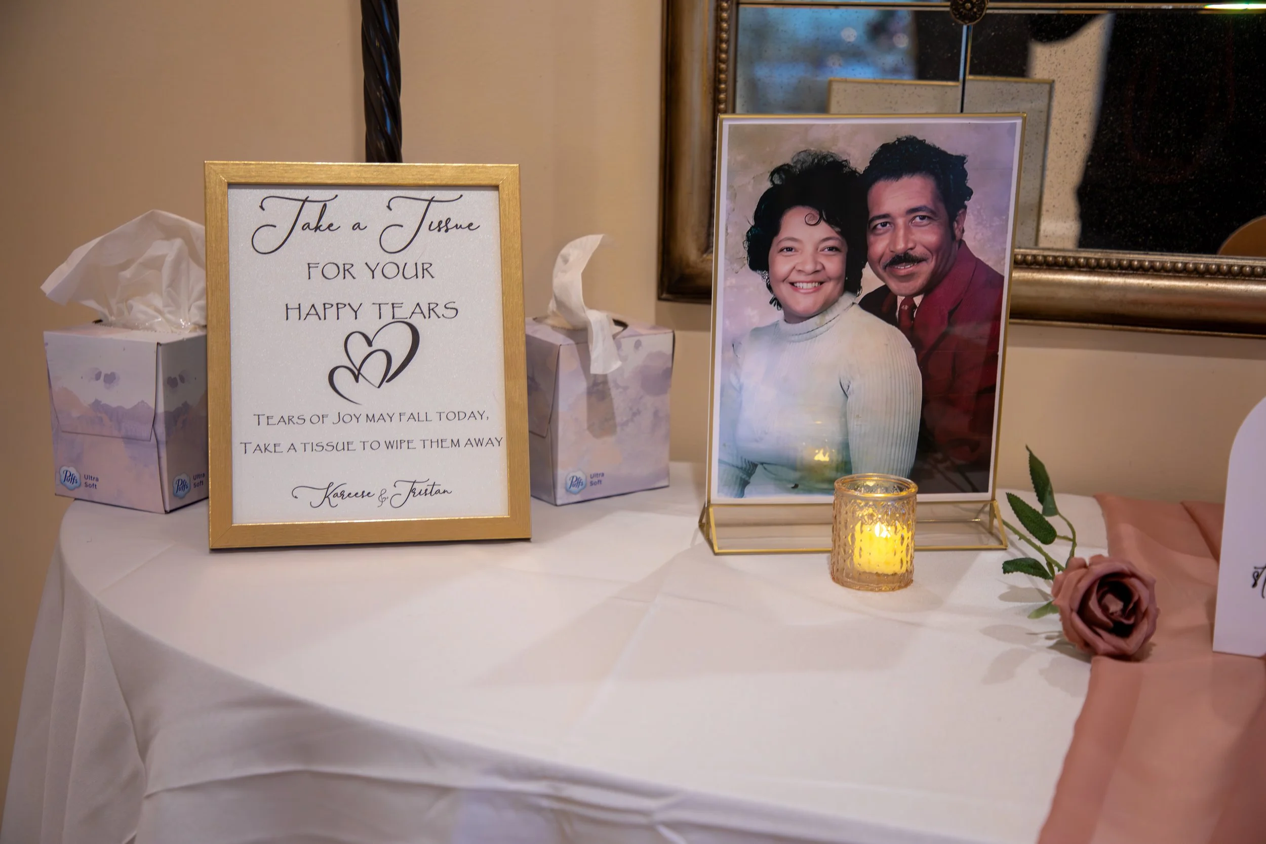 Wedding Photography by Will Locke. Table with tissues, a framed note, a framed wedding photo of a smiling couple, a lit candle, and a pink artificial rose.