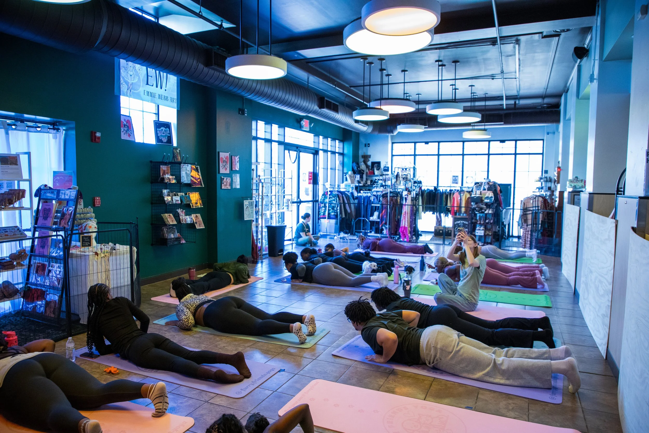 People participating in a yoga class inside a brightly lit retail store, with diverse attendees lying on yoga mats in a plank pose, instructor seated on the floor. Marketing and Event Photography by Will Locke. 