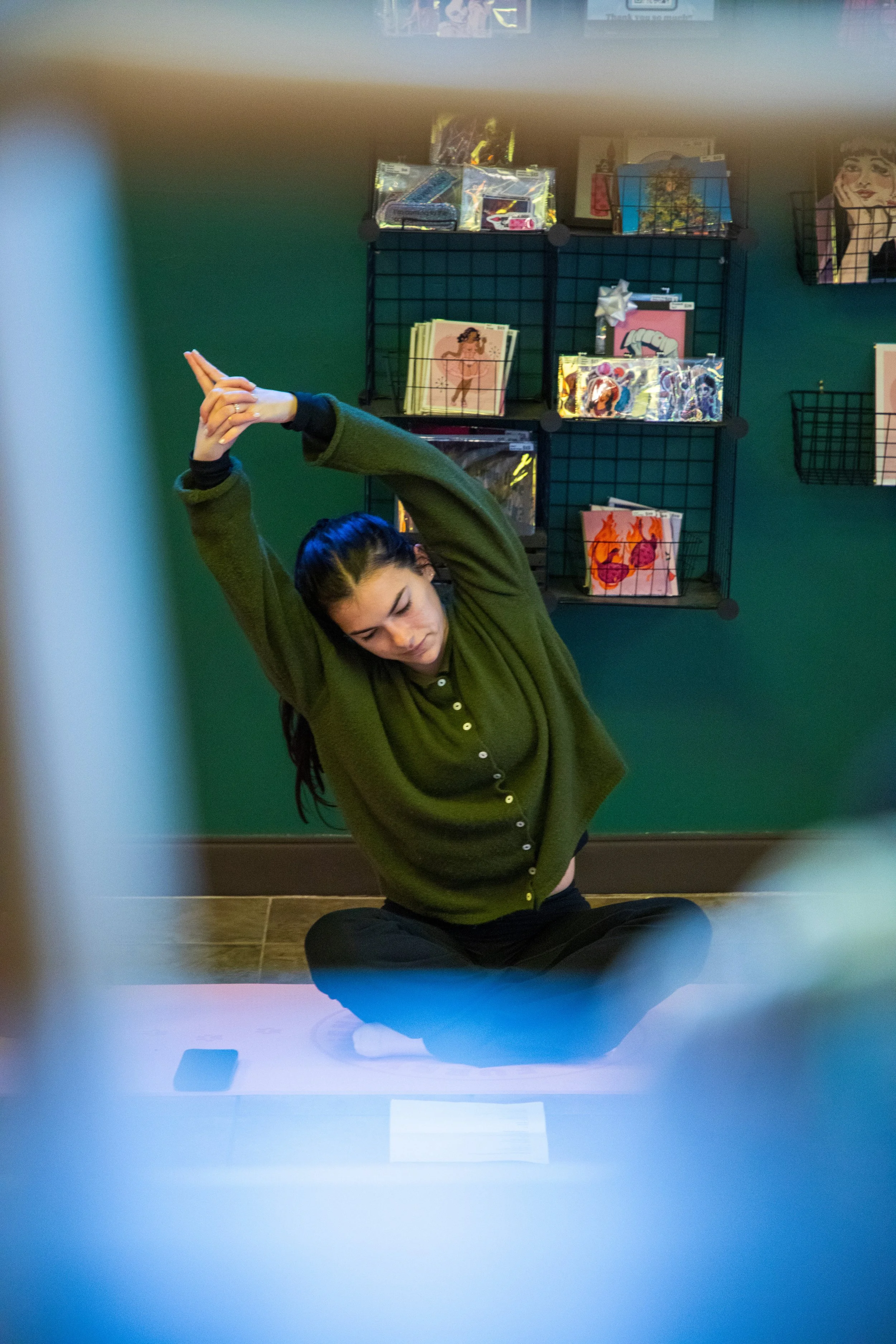 Marketing and Event Photography by Will Locke. A woman sitting on the floor stretching with her arms raised above her head in a room with green walls, decorated with art and merchandise on wire shelves.