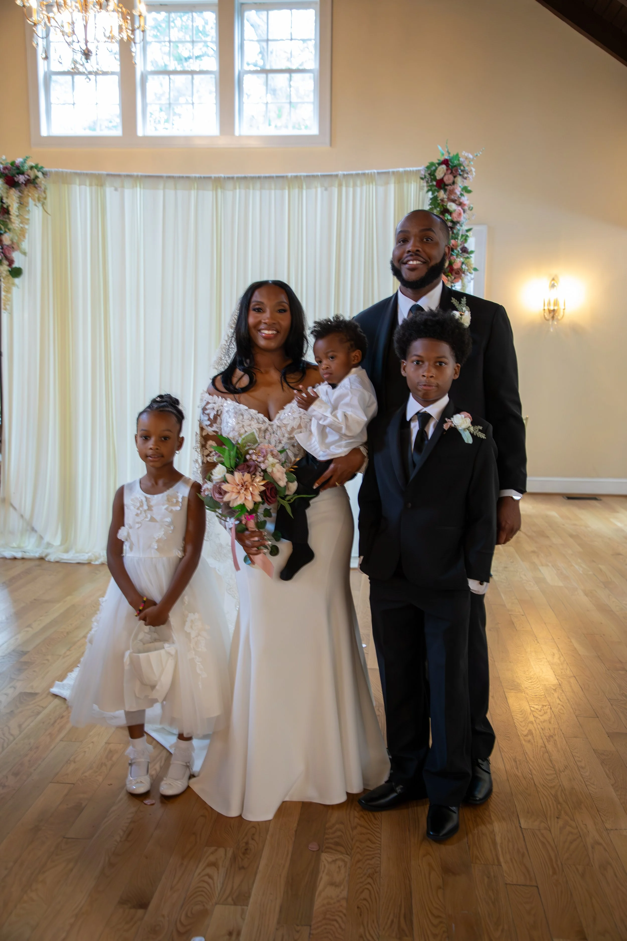 A family is dressed in formal wedding attire, standing indoors with a decorated backdrop of light-colored drapes and floral arrangements. Photo & Video by Will Locke Wedding Photography at the Woman's Club of Portsmouth in VA. 