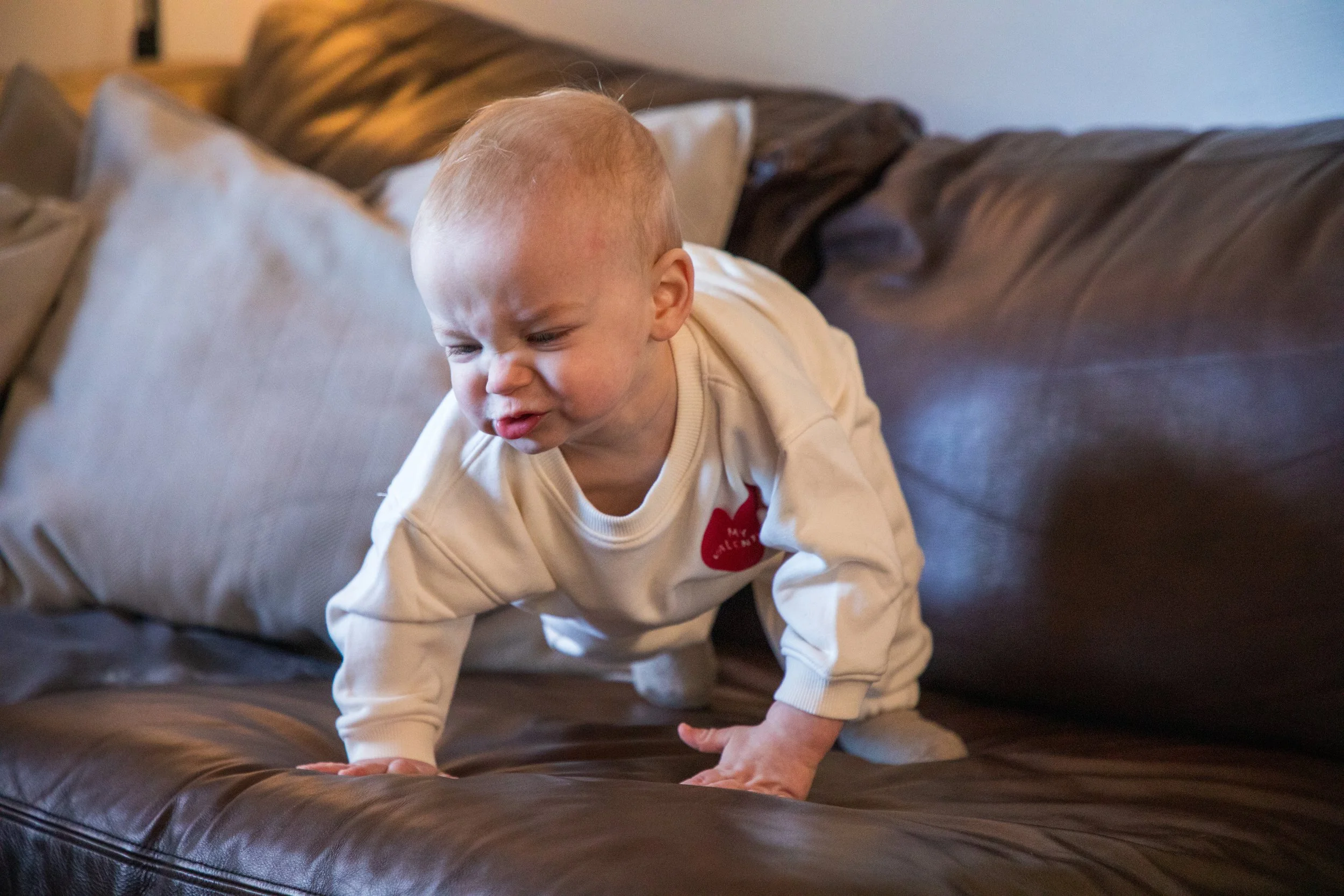 Baby Photography by Will Locke. A baby crawling on a dark brown leather couch, making a grimacing facial expression, wearing a cream-colored long-sleeve shirt with a red apple graphic.