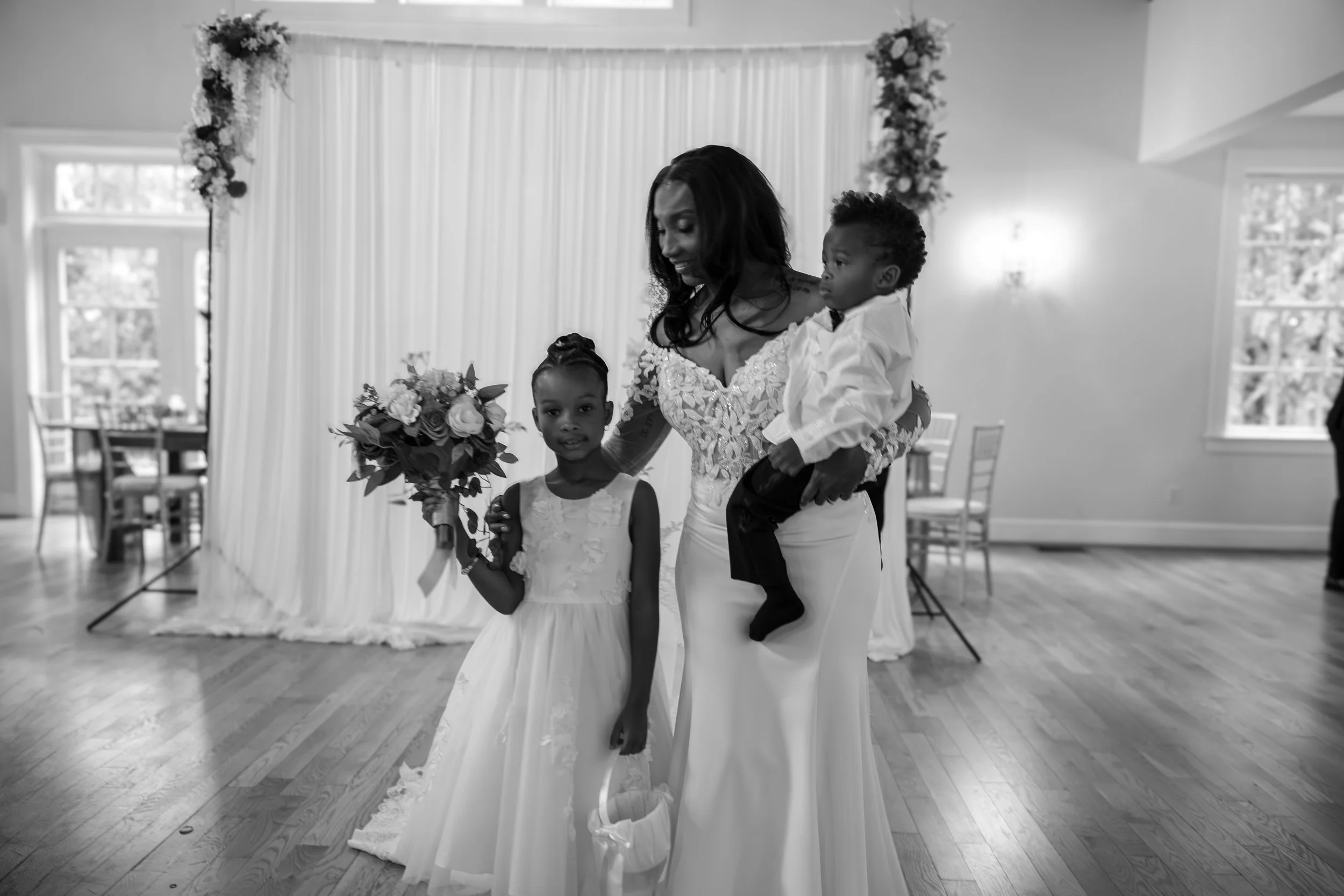 Photo & Video by Will Locke Wedding Photography at the Woman's Club of Portsmouth in Virginia. A woman in a wedding dress holding two young children, one girl holding a bouquet and one boy being held, in a decorated indoor venue.